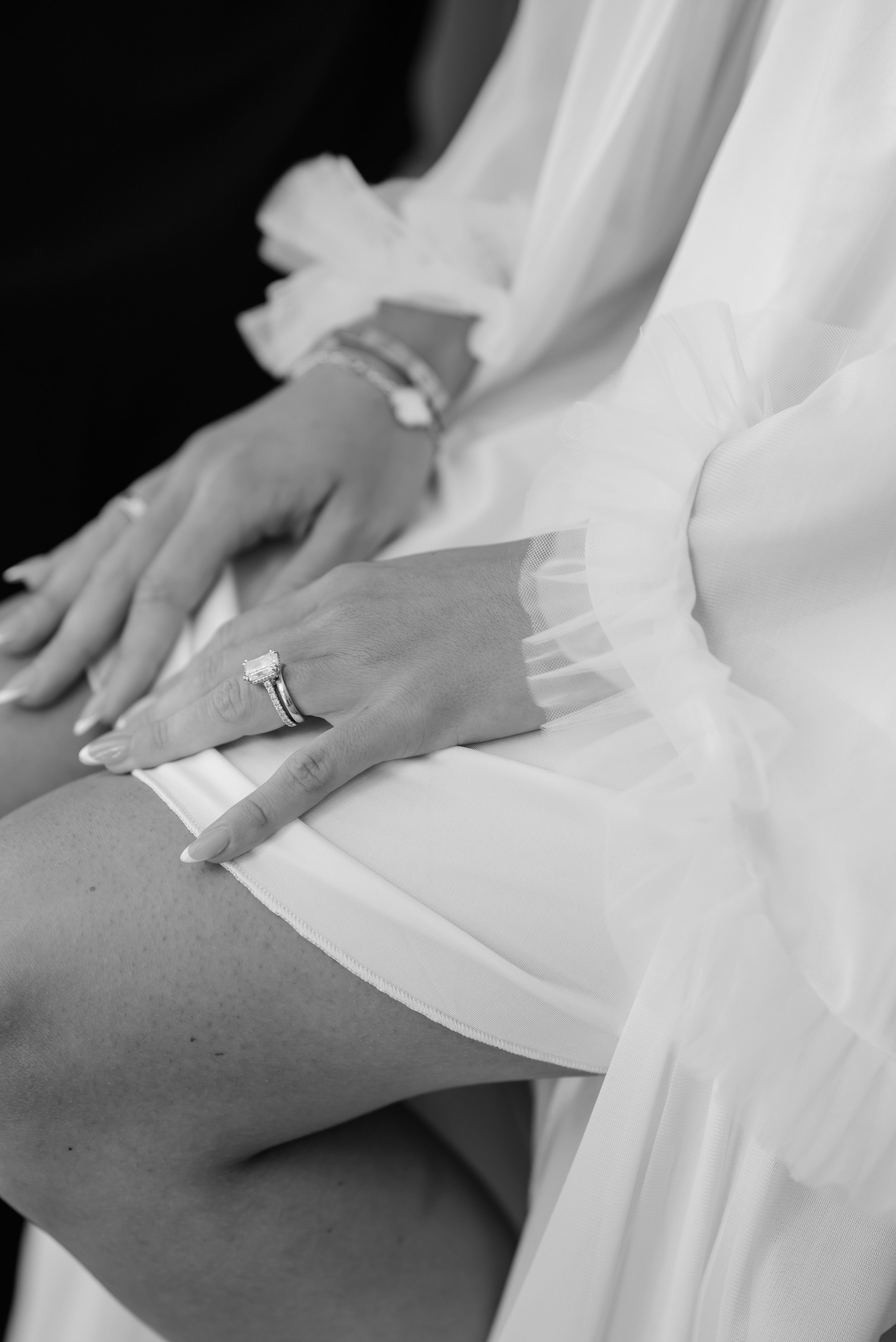Close-up of a woman's hands resting on her lap, wearing a large ring and a bracelet, with her fingers extended over her thigh. She is dressed in a white garment with tulle or sheer fabric details.