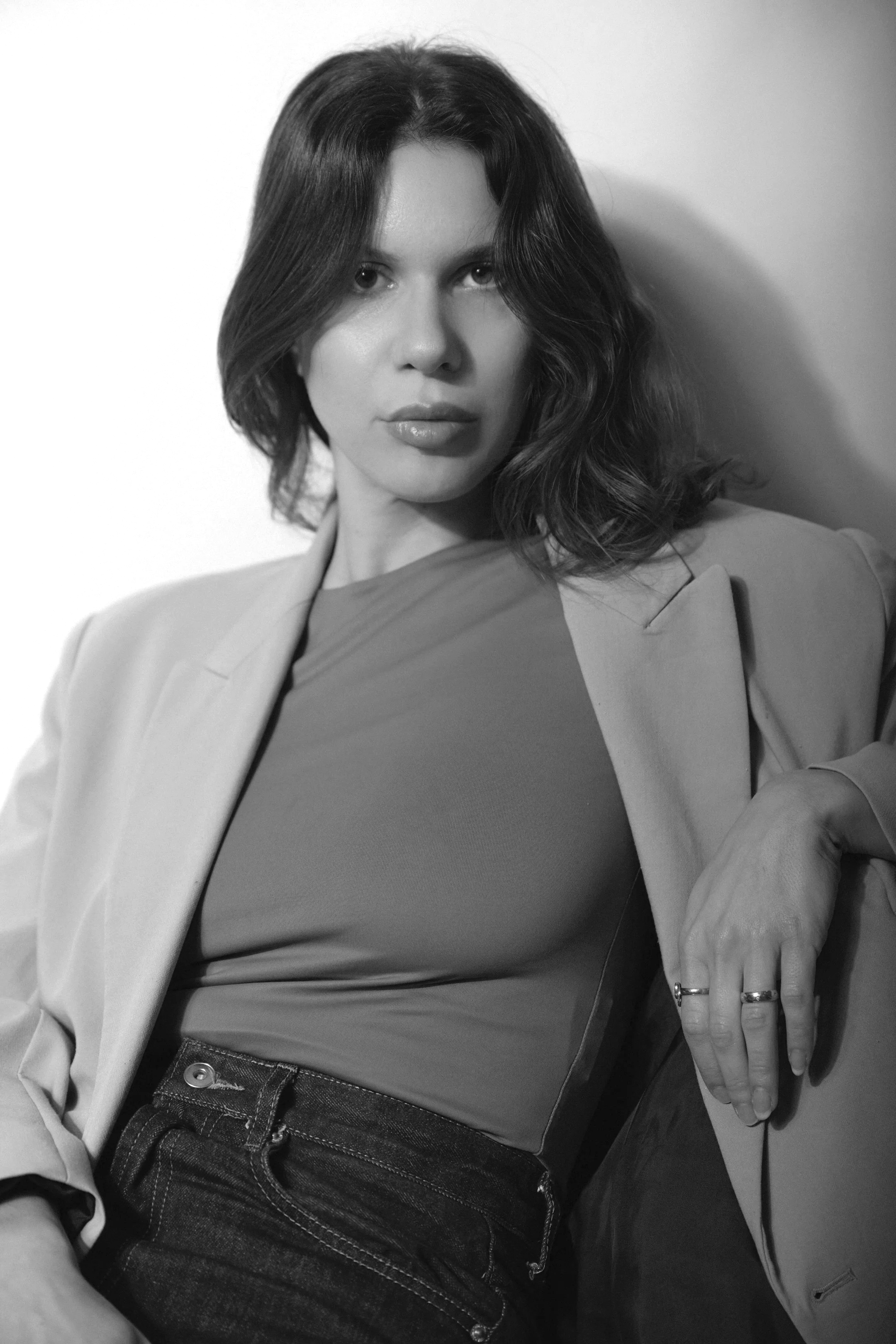 Black and white photo of a woman with shoulder-length wavy hair, wearing a blazer over a t-shirt, leaning against a wall with her left hand resting on her knee.