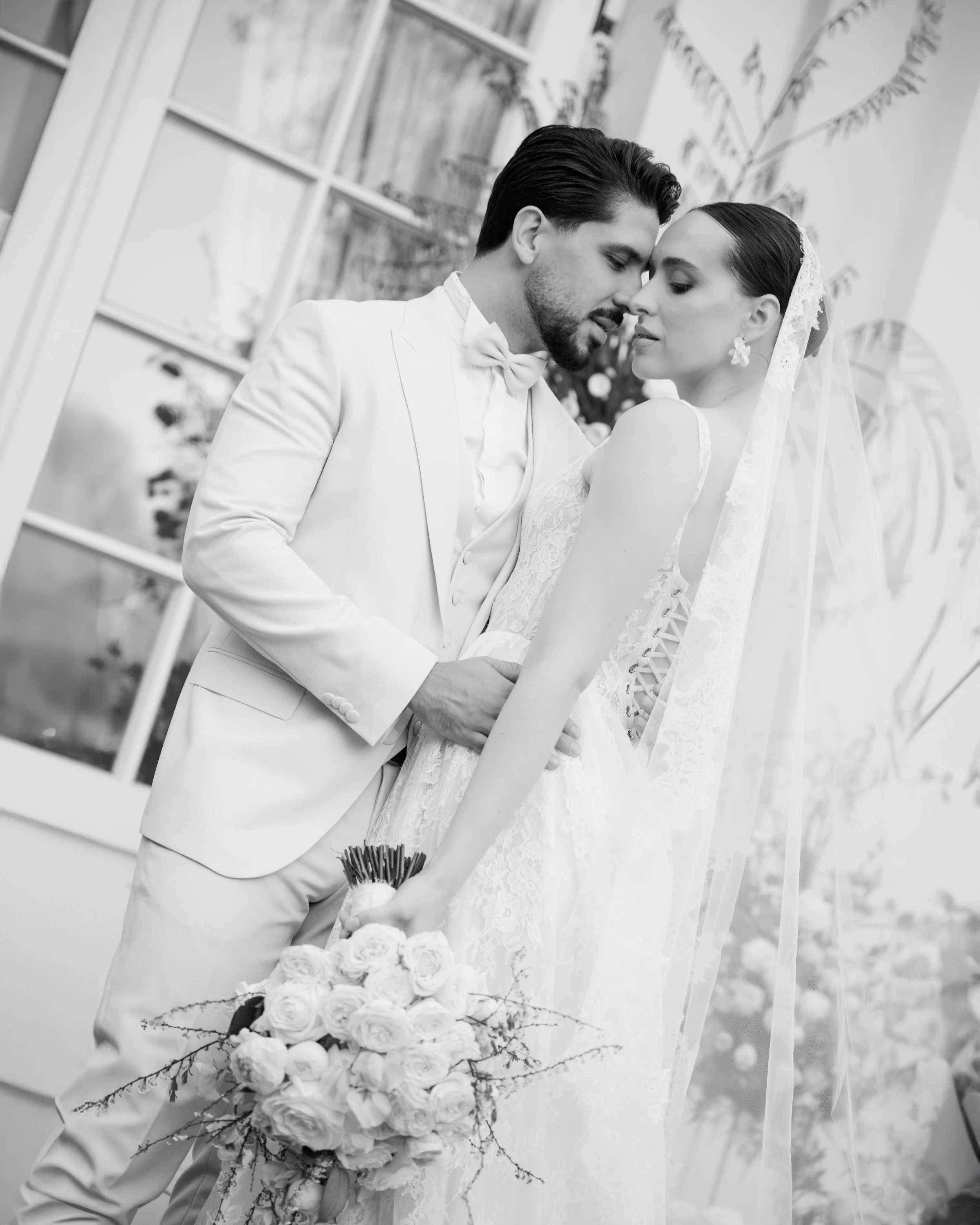 Black and white photo of a bride and groom in an intimate pose, with their foreheads touching and eyes closed. The groom is dressed in a white suit with a bow tie, and the bride in a lace wedding dress holding a bouquet of roses. They are indoors with a large window and houseplants in the background.