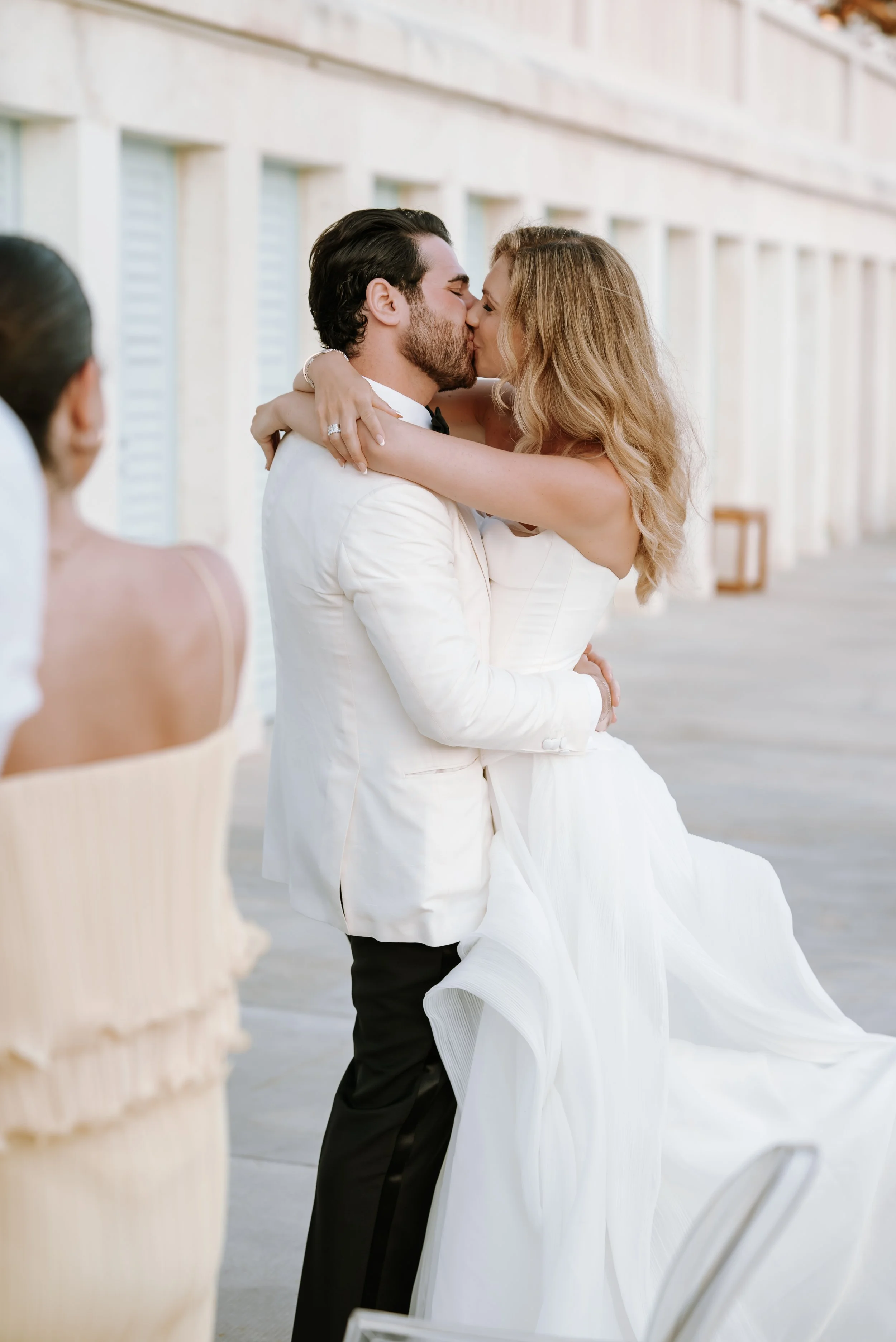 A bride and groom share a kiss at their wedding reception, with the groom lifting the bride in a white wedding gown and the bride in a strapless white dress, in an outdoor setting with a building in the background. RebornWeddings Barbara Jerković