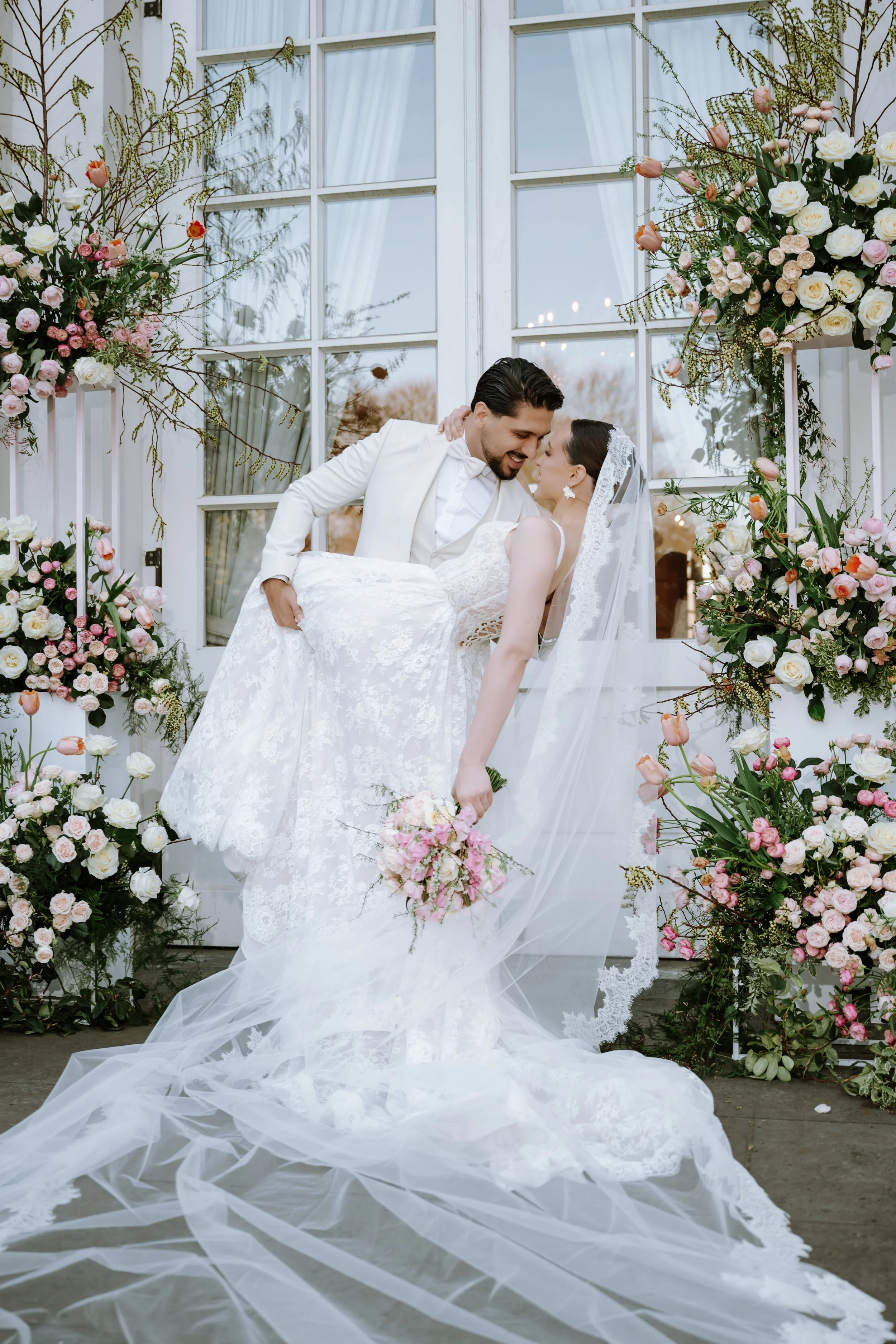 A bride and groom sharing a dance at their wedding, with the groom lifting the bride. The bride is holding a bouquet and wearing a lace wedding gown and veil. They are surrounded by floral arrangements with pink and white flowers, in front of a large window with white framed panes.