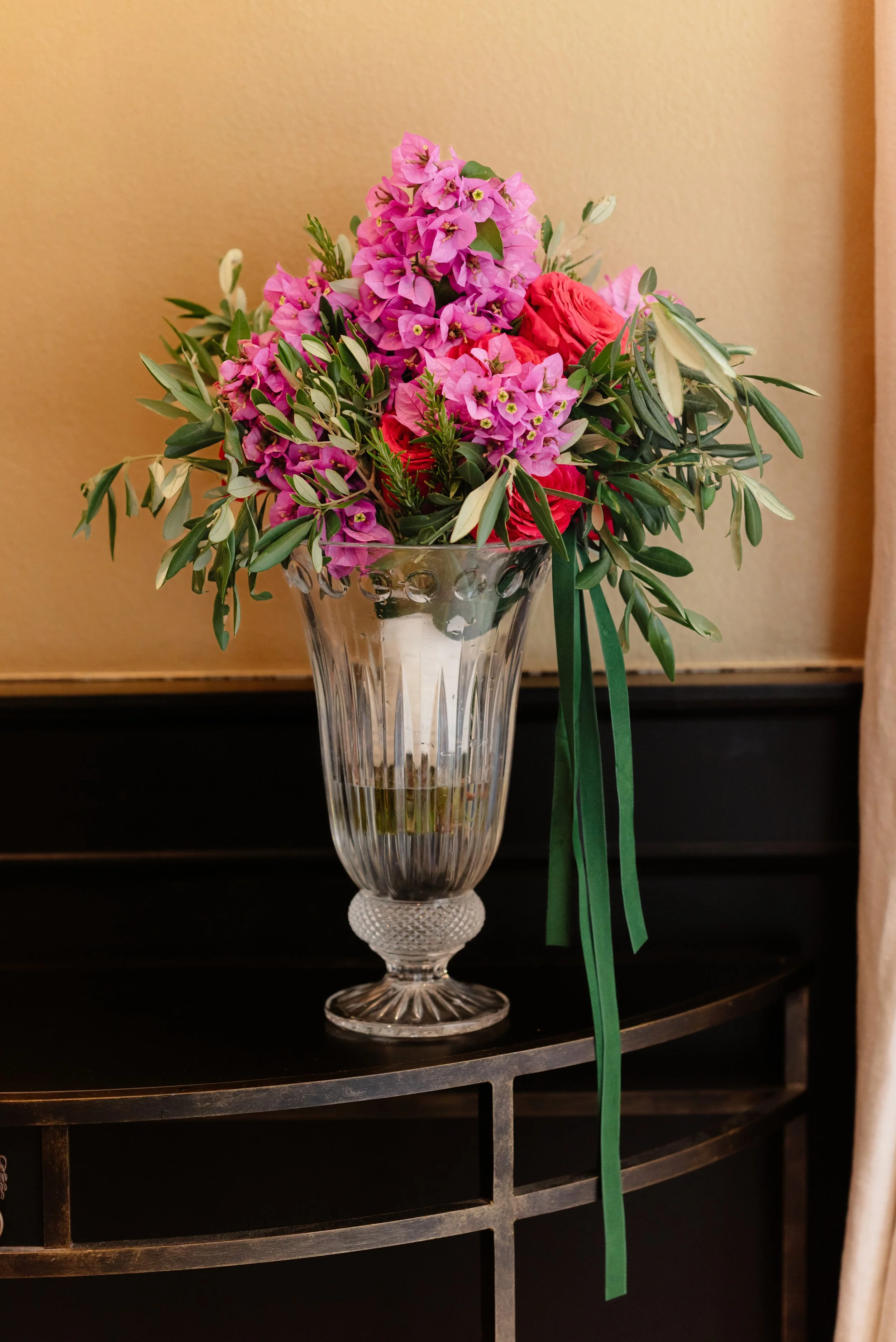 A tall, clear glass vase filled with pink and red flowers, including pink bougainvillea and red roses, with green foliage, placed on a black wooden table against a beige wall. RebornWeddings Barbara Jerković Golob Hvar Wedding Photographer