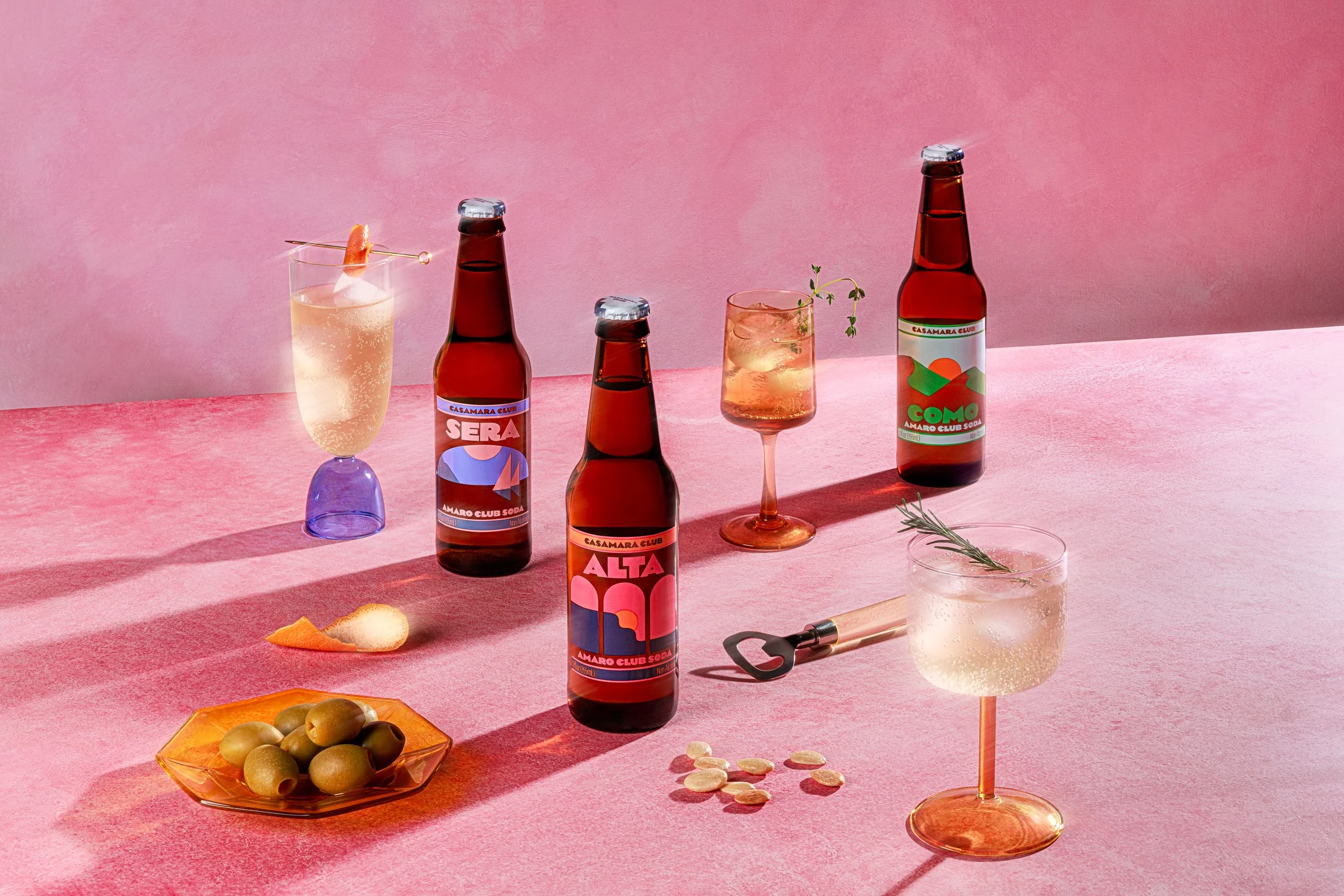 Two brown glass bottles with colorful labels, one glass containing a pink drink with ice and a sprig of greenery, a smaller glass with a similar drink and greenery, another glass with a bubbly light-colored beverage and ice, a plate of green olives, a corkscrew, and some sliced almonds, all on a pink surface with a pink background.