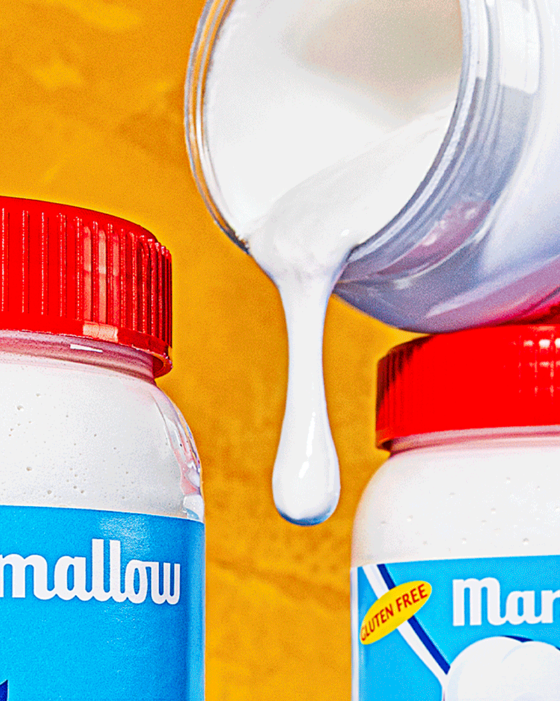 A container of white liquid, likely milk, pouring into two bottles of Mallow marshmallow snack ingredients with red caps on a yellow background.