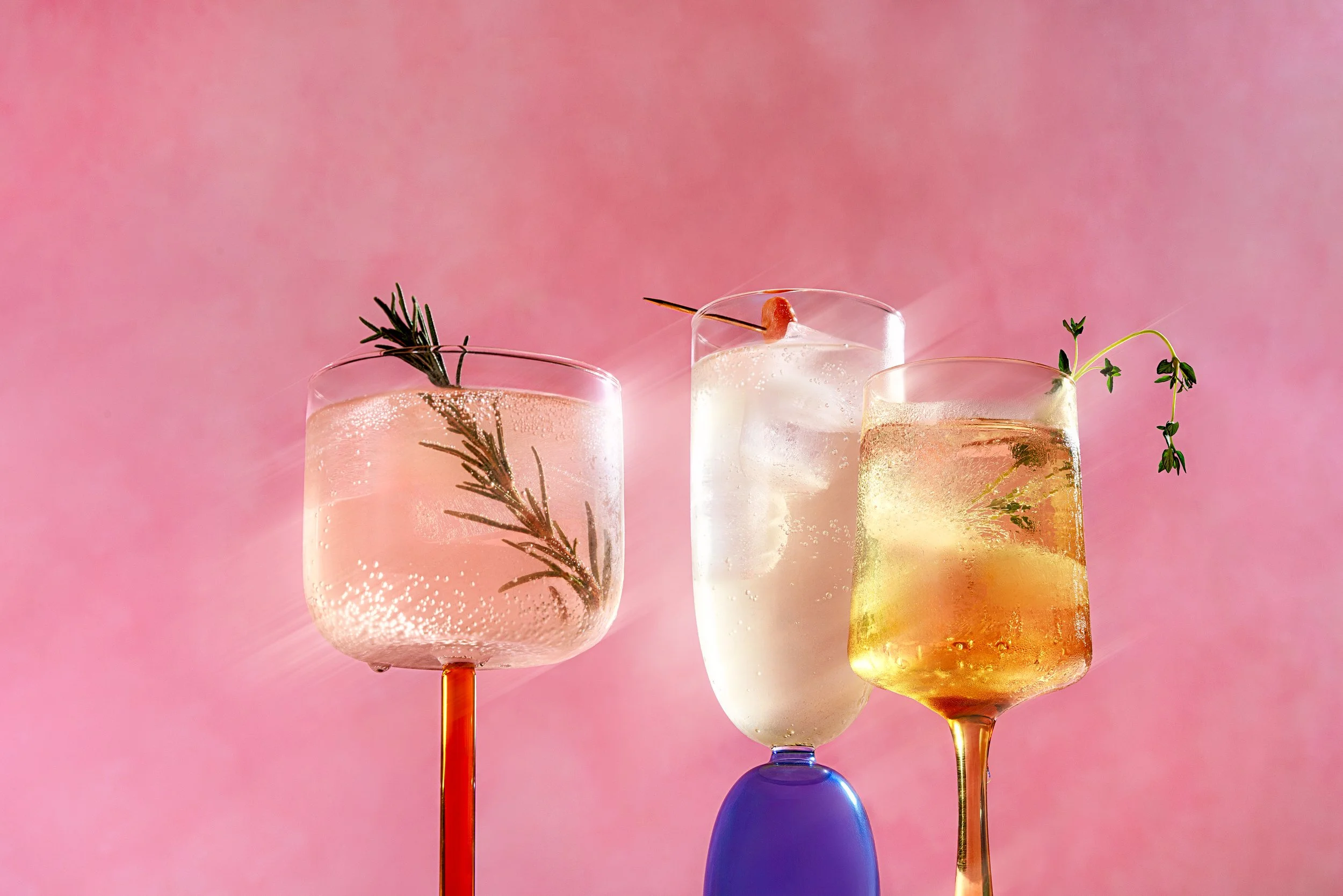 Four hanging glasses with colorful drinks, garnished with herbs and olives, against a pink background.