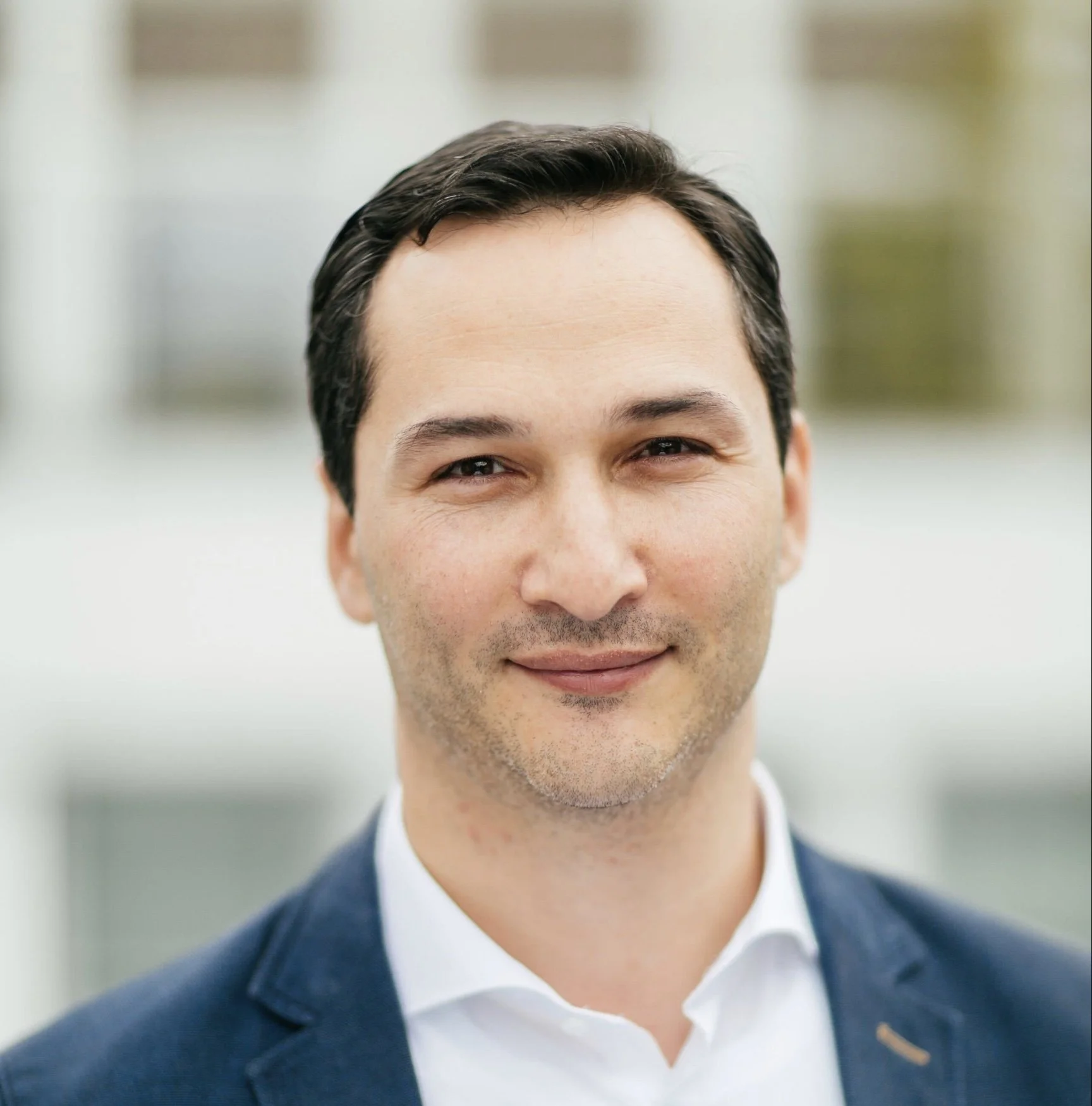 Close-up of a smiling man wearing a suit outside with a blurred building background.