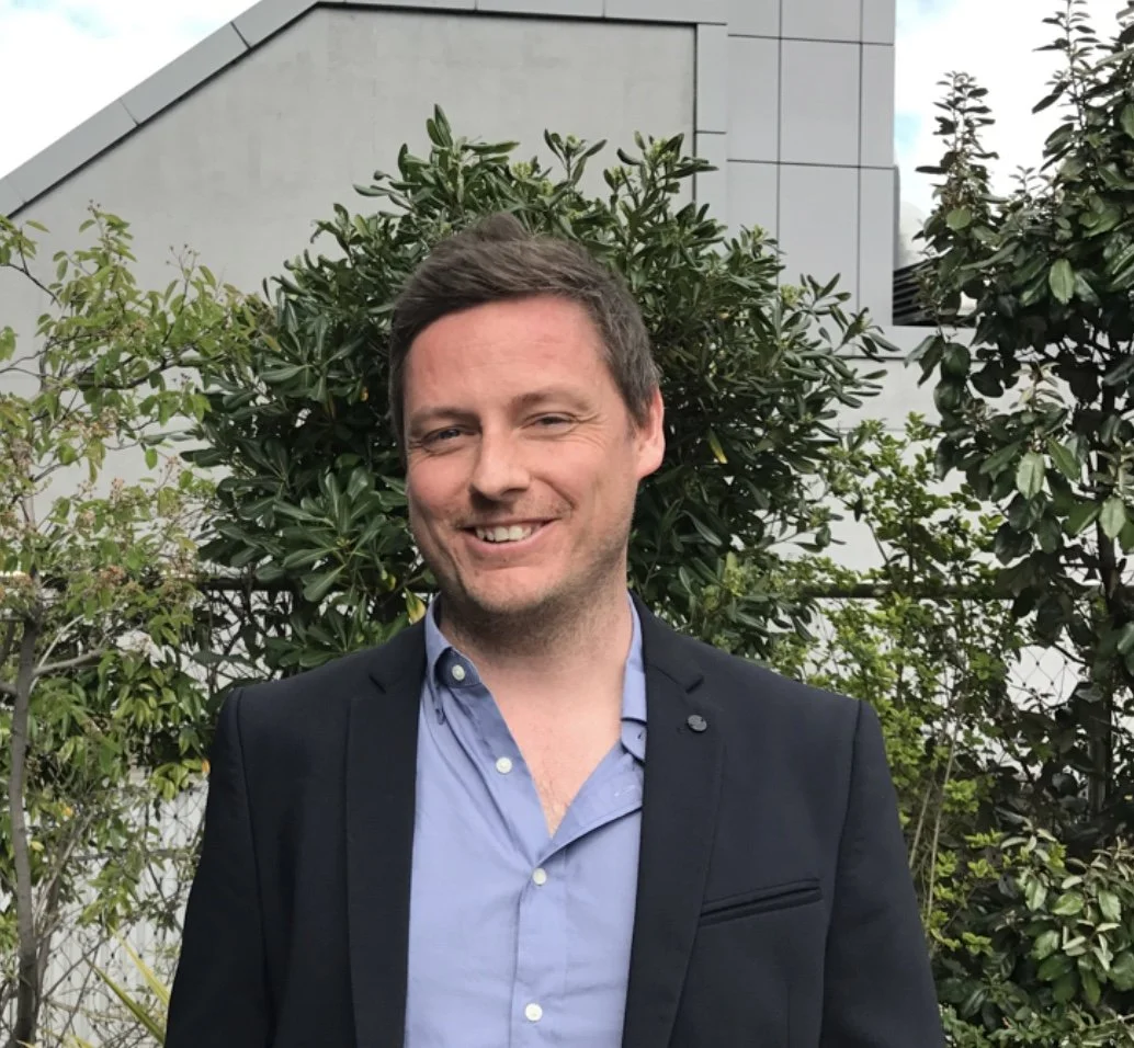 A man in a black blazer and light blue shirt smiling outdoors, standing in front of green shrubbery and trees with a modern building in the background.