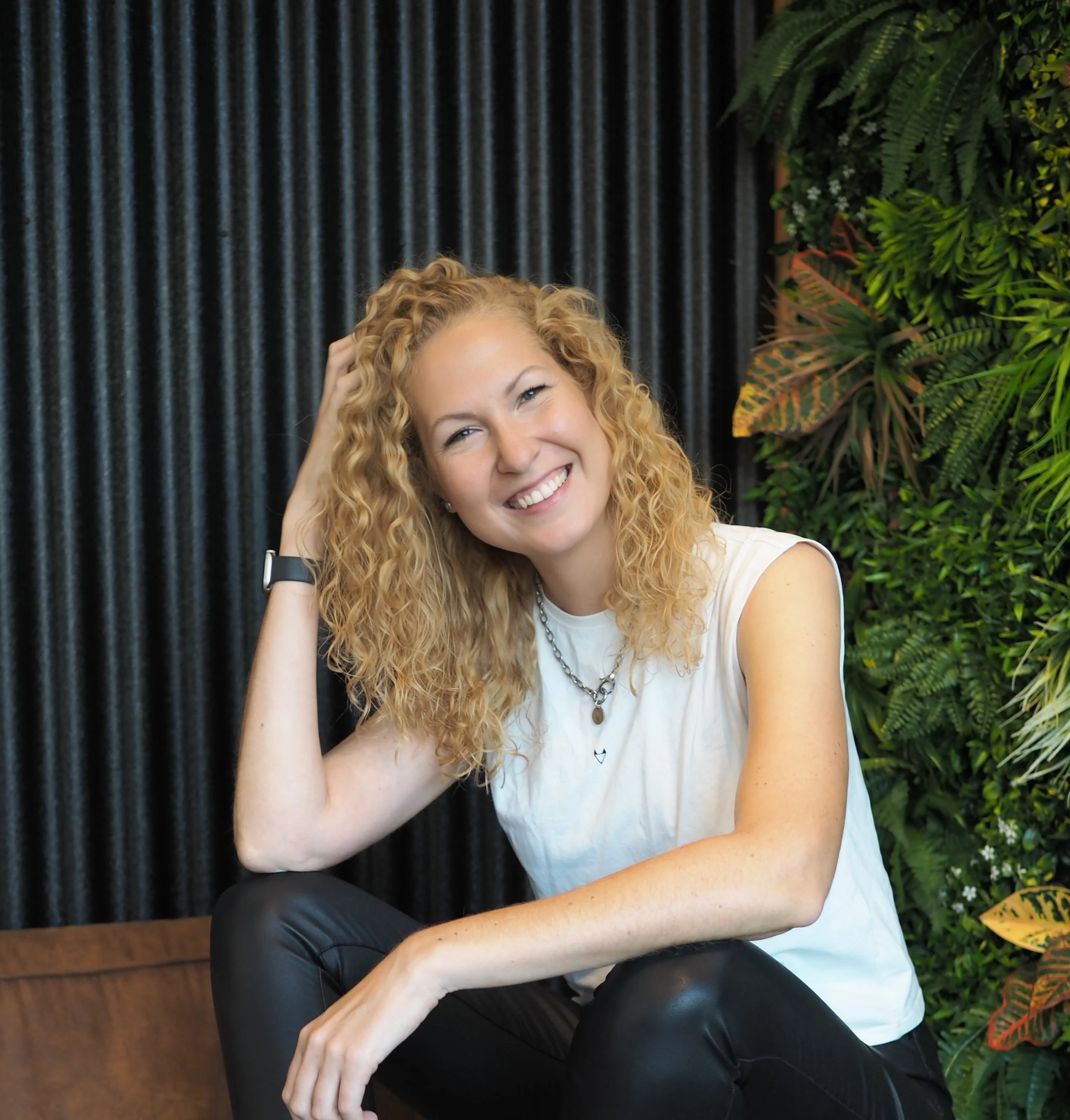 A young woman with curly blonde hair smiling and sitting against a black curtain and green foliage background.