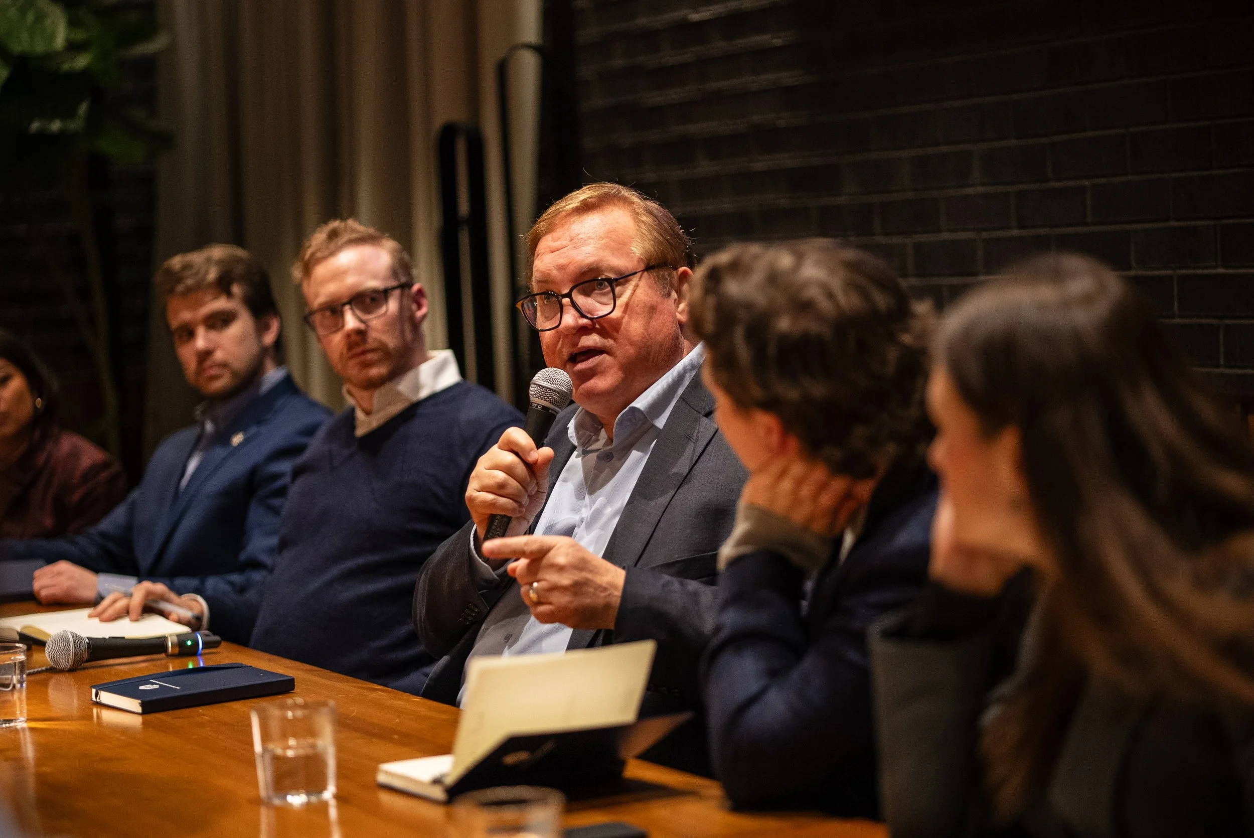 Man in glasses holding a microphone speaking at a panel discussion with others sitting beside him at a wooden table.