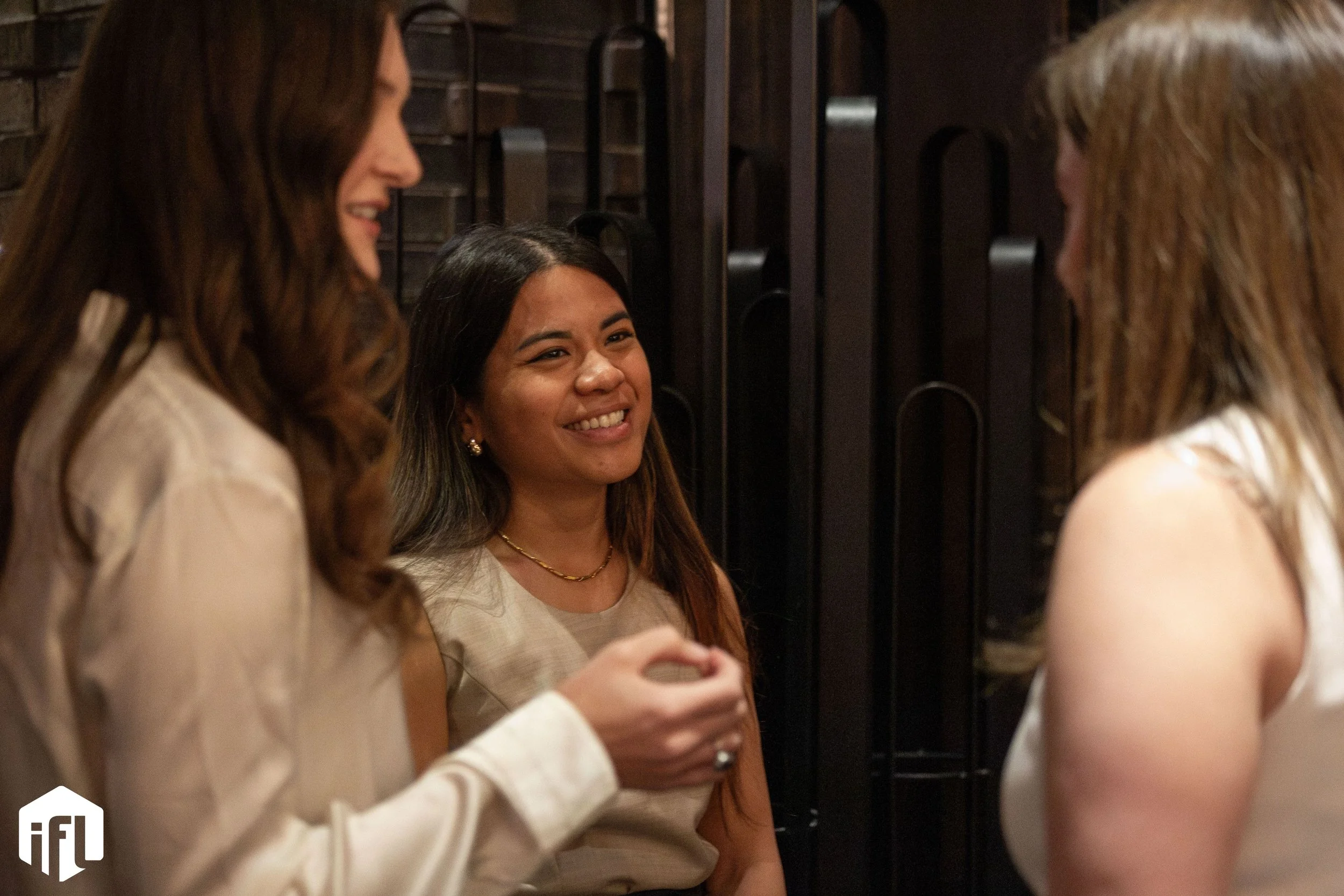 Three women talking and smiling indoors, with a dark wall and decorative elements in the background.