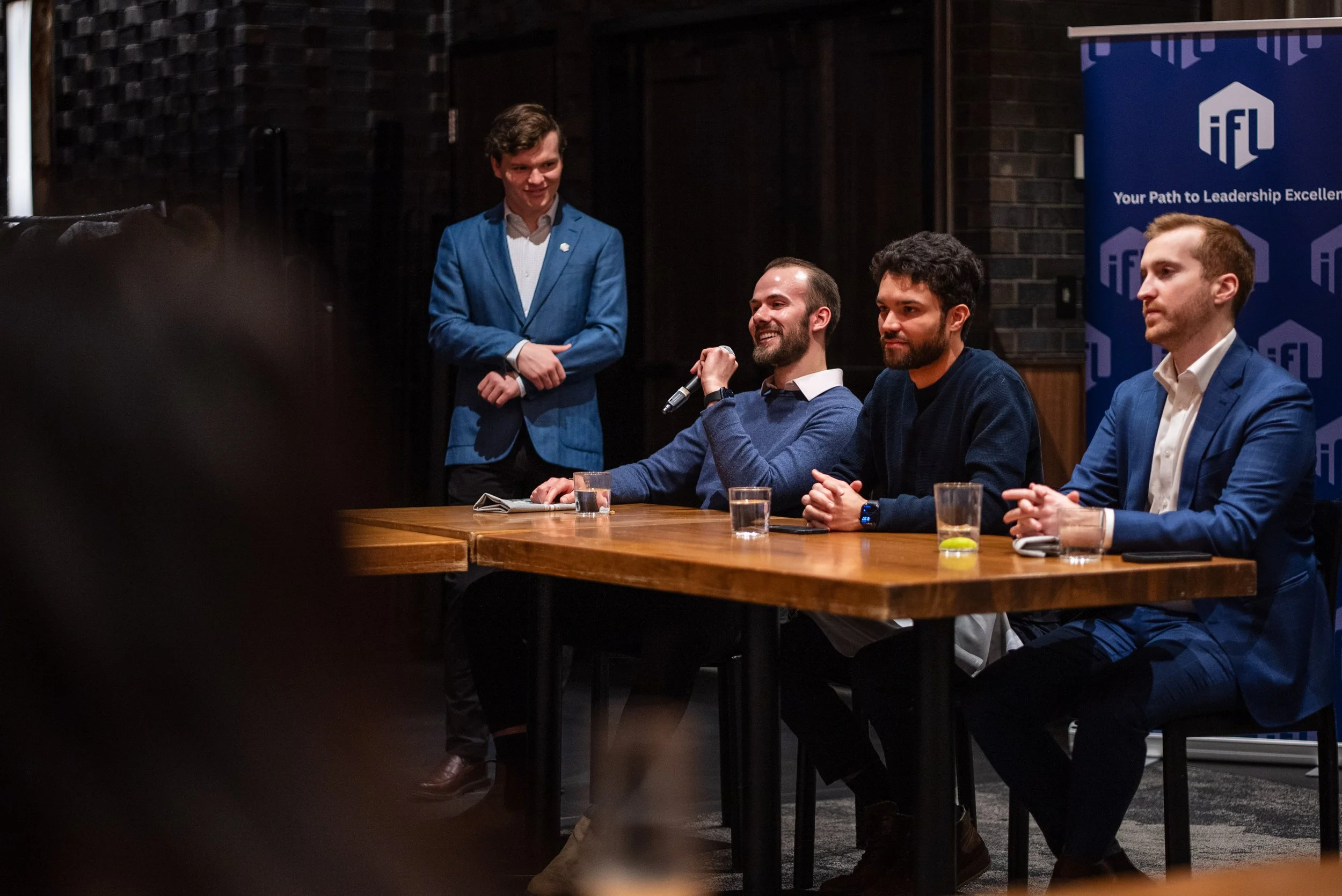 Four men in suits sitting at a conference table with drinks, one man speaking into a microphone, and a man standing in a blue suit in the background near a blue banner with the IFL logo.