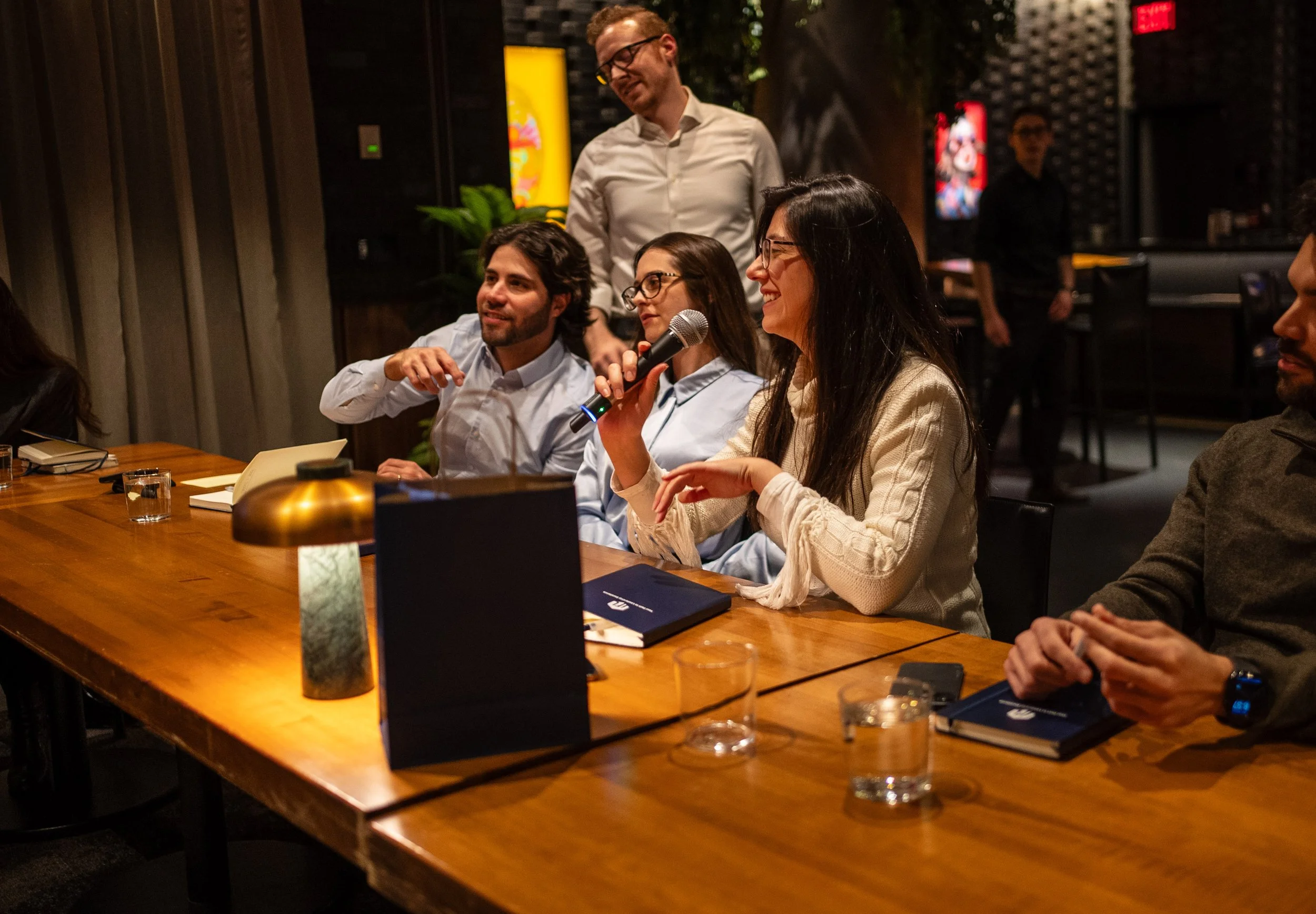 People sitting at a conference table, with one woman speaking into a microphone, in a dimly lit room with a man standing behind her and others listening.