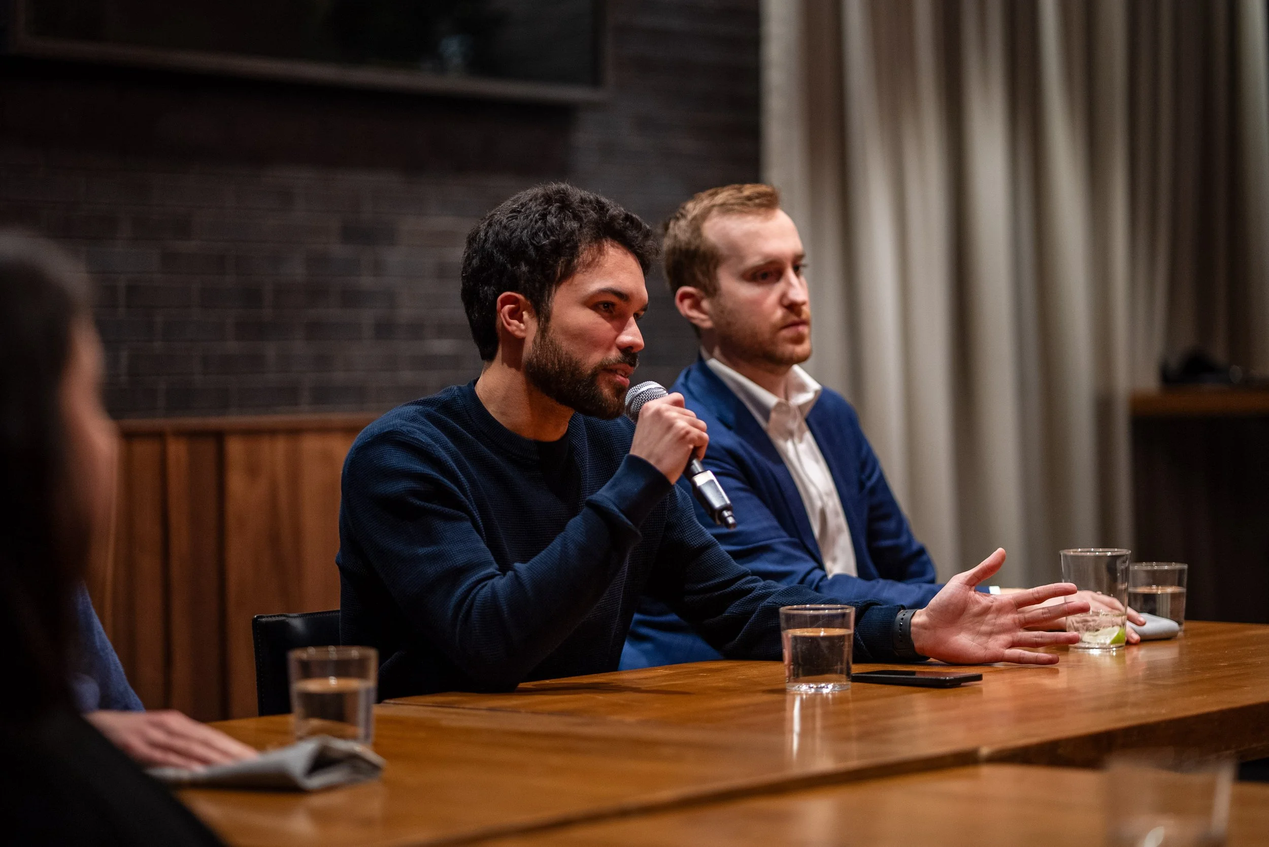 Two men sitting at a wooden table during a panel discussion or interview; one is speaking into a microphone, while the other listens; glasses of water are in front of them; background features a brick wall and curtains.