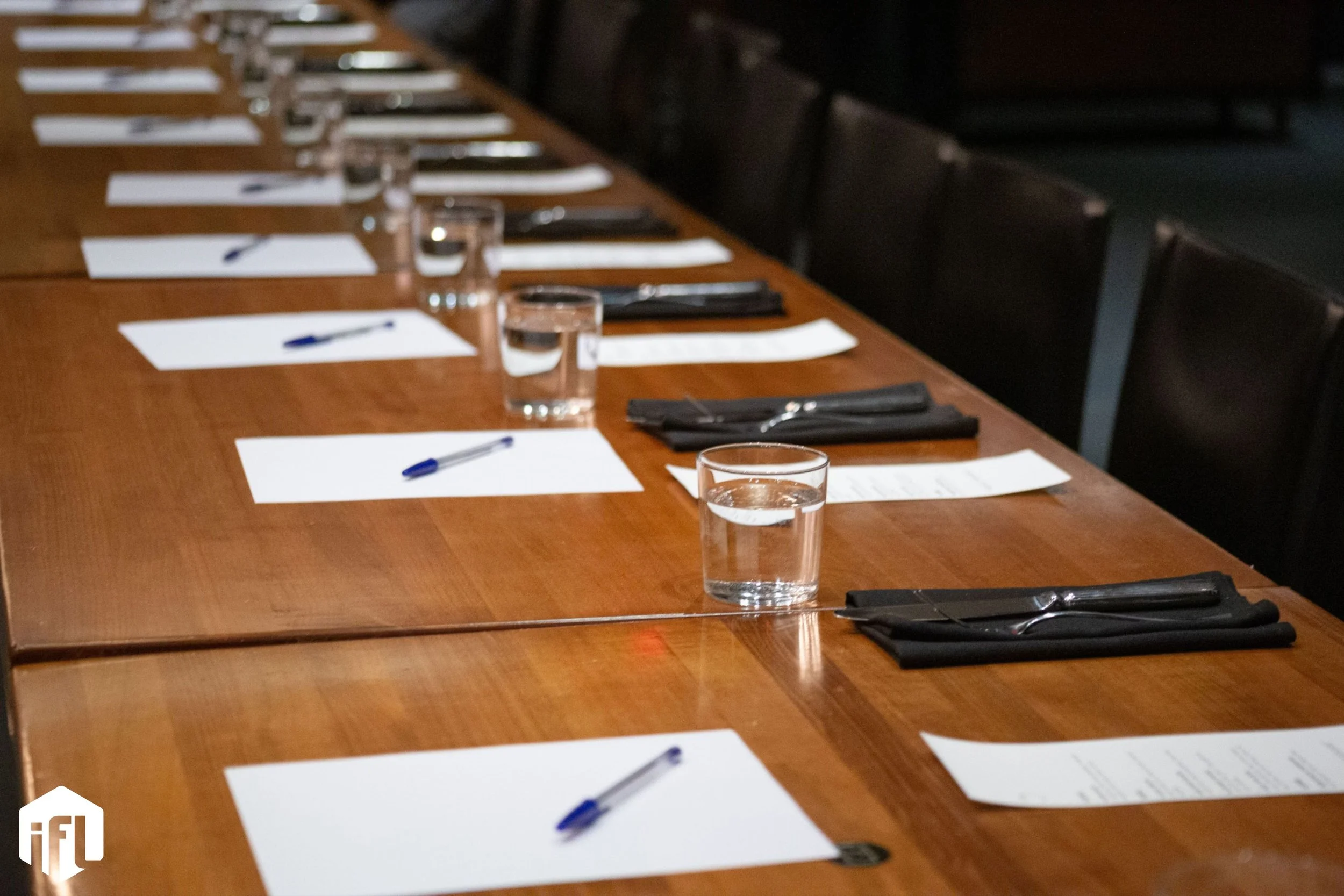 A long wooden table set with glasses of water, white notepads, blue pens, and black napkins for an event or meeting.