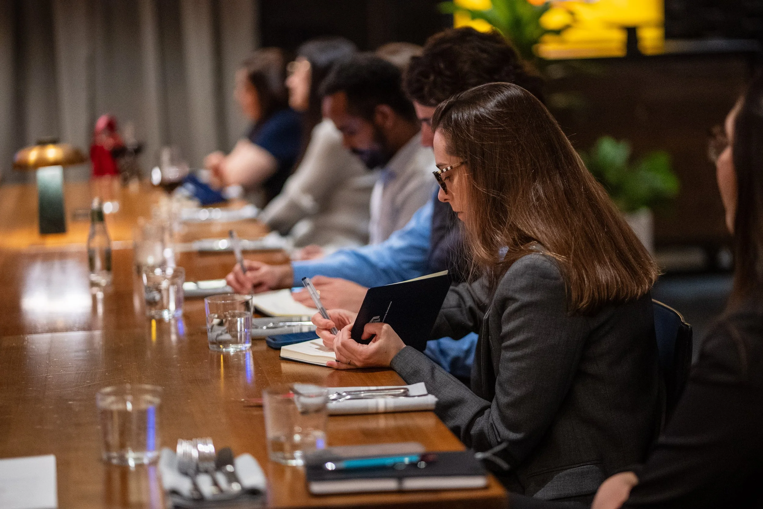 A group of people sitting at a long conference table, taking notes, with notebooks and glasses of water in front of them.