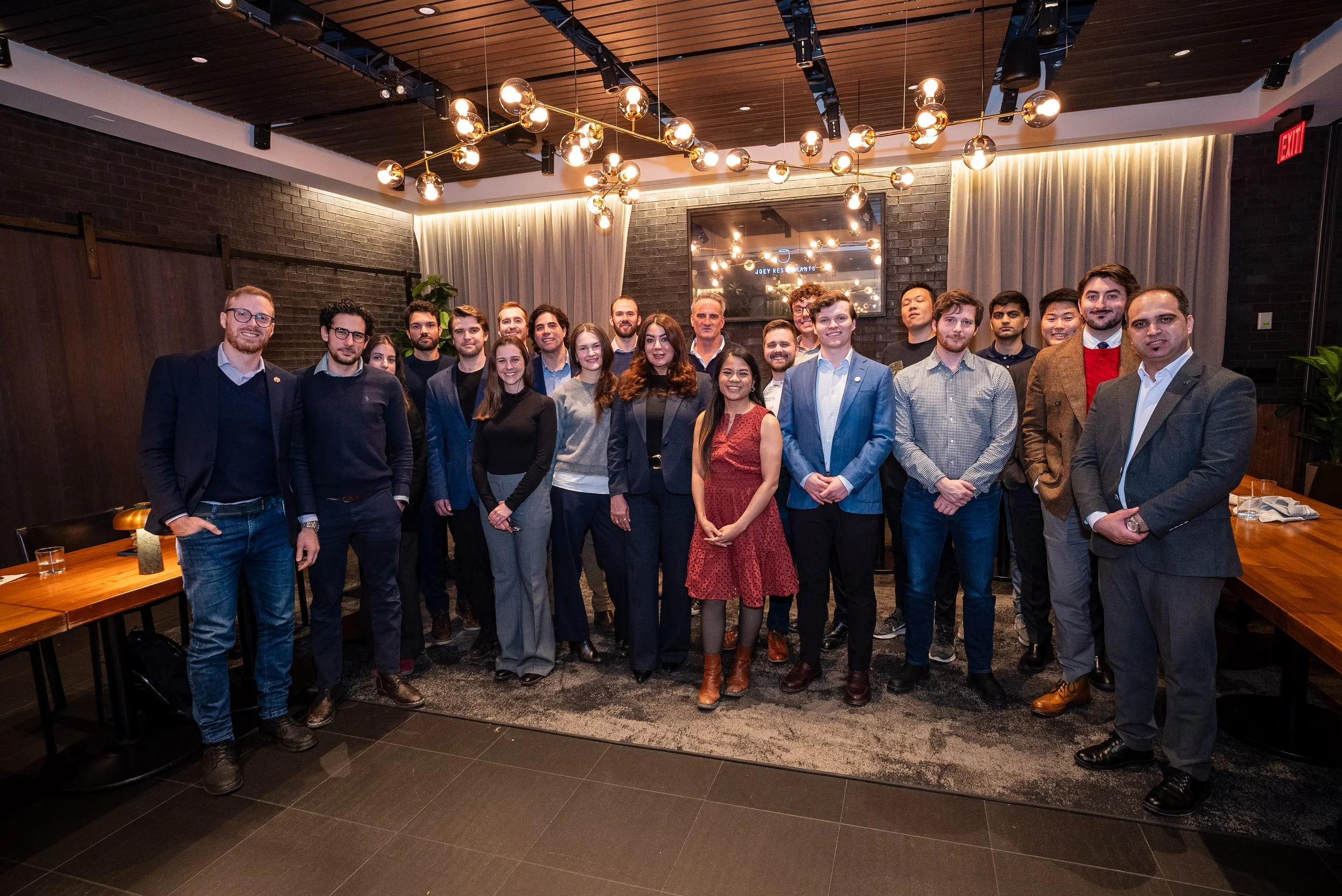 Group of professionally dressed people posing for a photo in a modern, stylish restaurant or event space with dark brick walls, warm lighting, and curtains in the background.