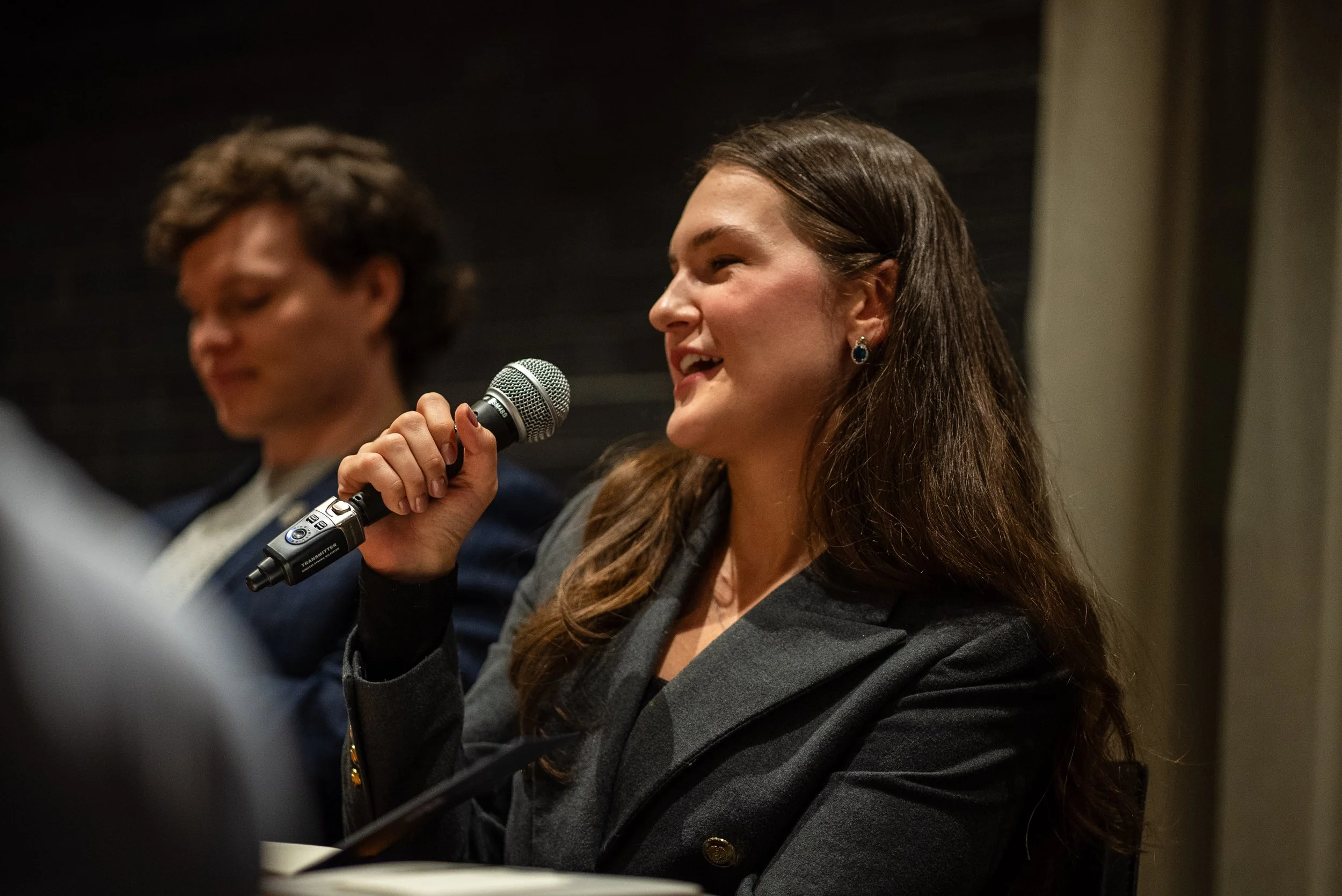 A woman with long brown hair speaking into a microphone during a panel or discussion, with a man with curly hair sitting beside her, blurred in the background.