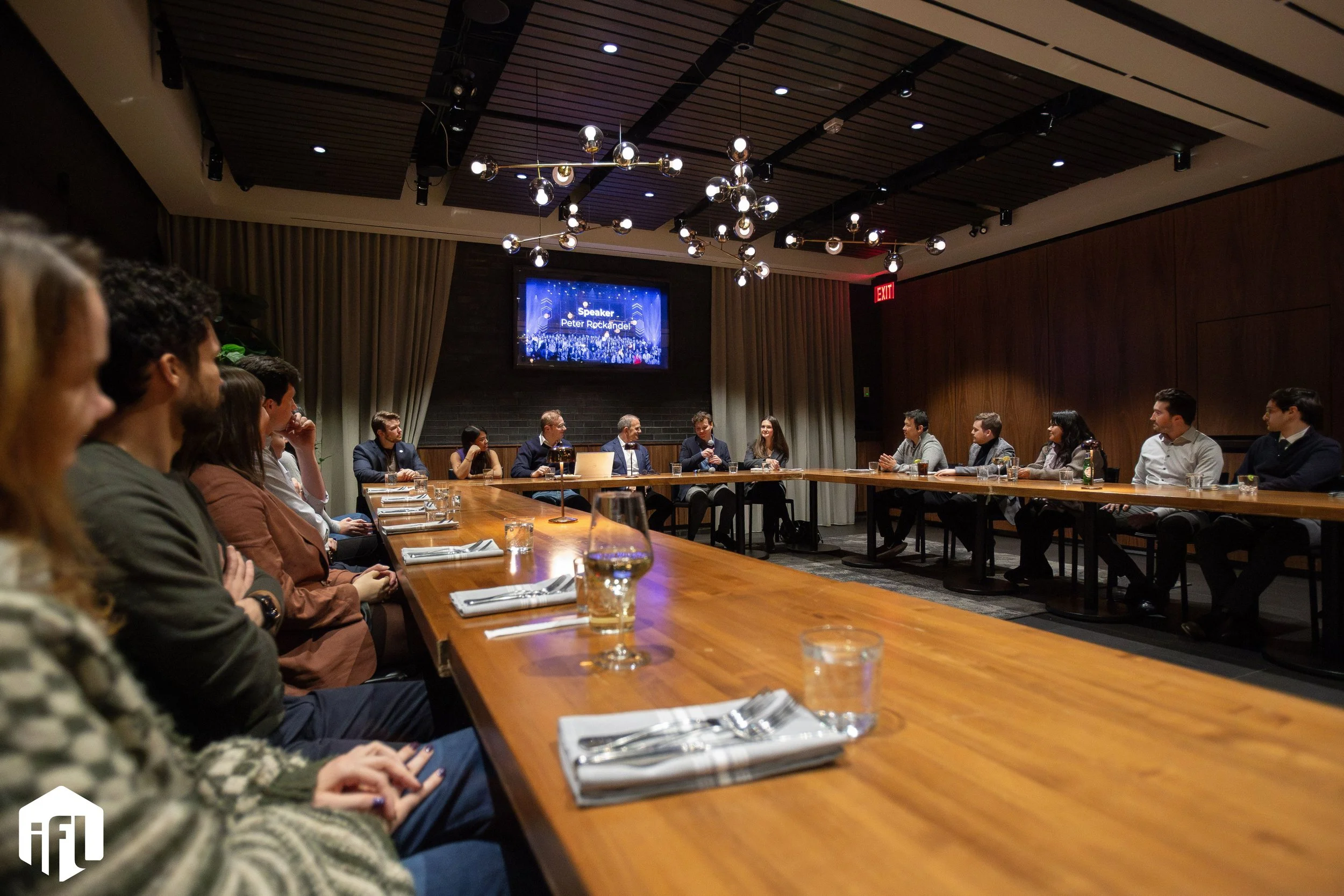 Business meeting or panel discussion with people seated around a large, U-shaped wooden table in a modern conference room with a dark wood-paneled wall, a large screen displaying the speaker's name, and ceiling lighting fixtures.