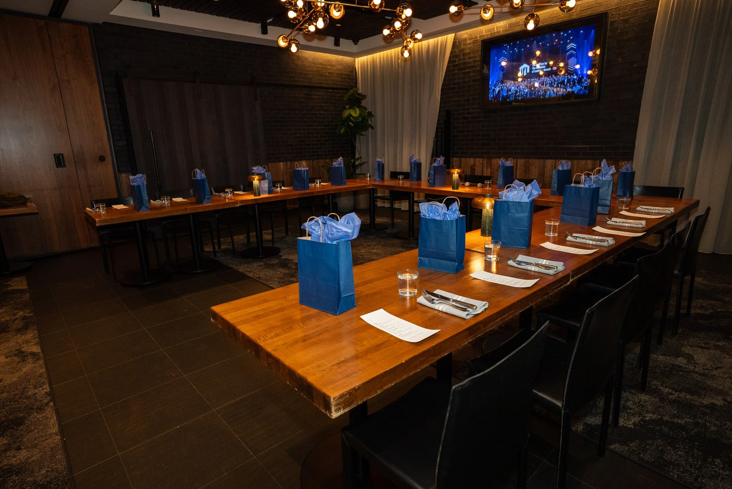 A conference or meeting room set up for an event with a U-shaped arrangement of wooden tables. Each place setting includes silverware, a napkin, a printed menu or program, a glass of water, and a blue gift bag with tissue paper. The room features dark flooring, brick and wood-paneled walls, a modern chandelier, and a wall-mounted flat-screen TV displaying a blue-themed event logo and audience.
