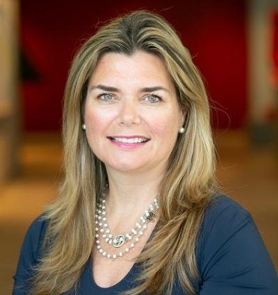 Portrait of a smiling woman with blonde hair, wearing a blue top and pearl jewelry, standing against a blurred indoor background.