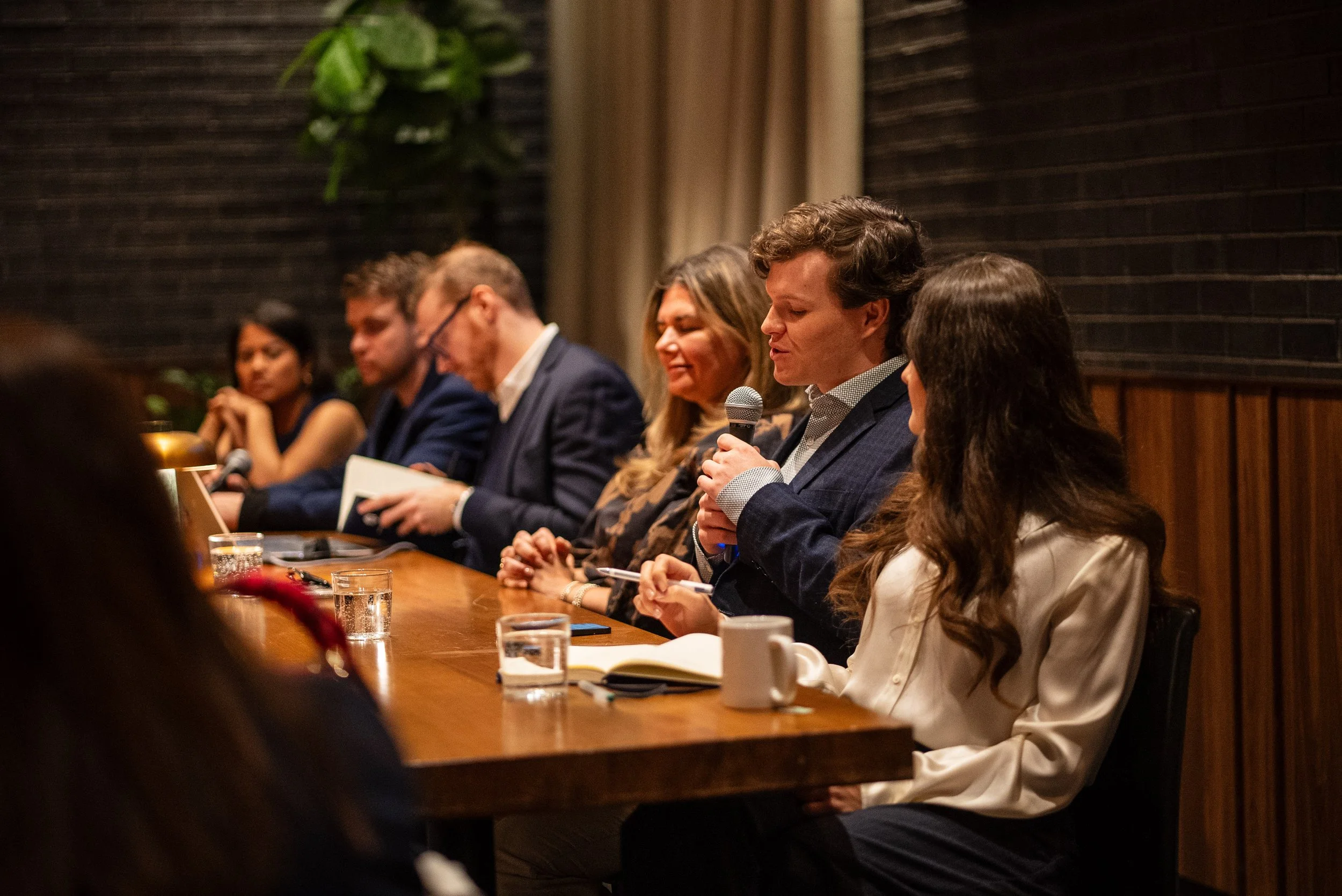 A man in a dark blazer speaking into a microphone at a panel discussion, surrounded by other people seated at a long table.