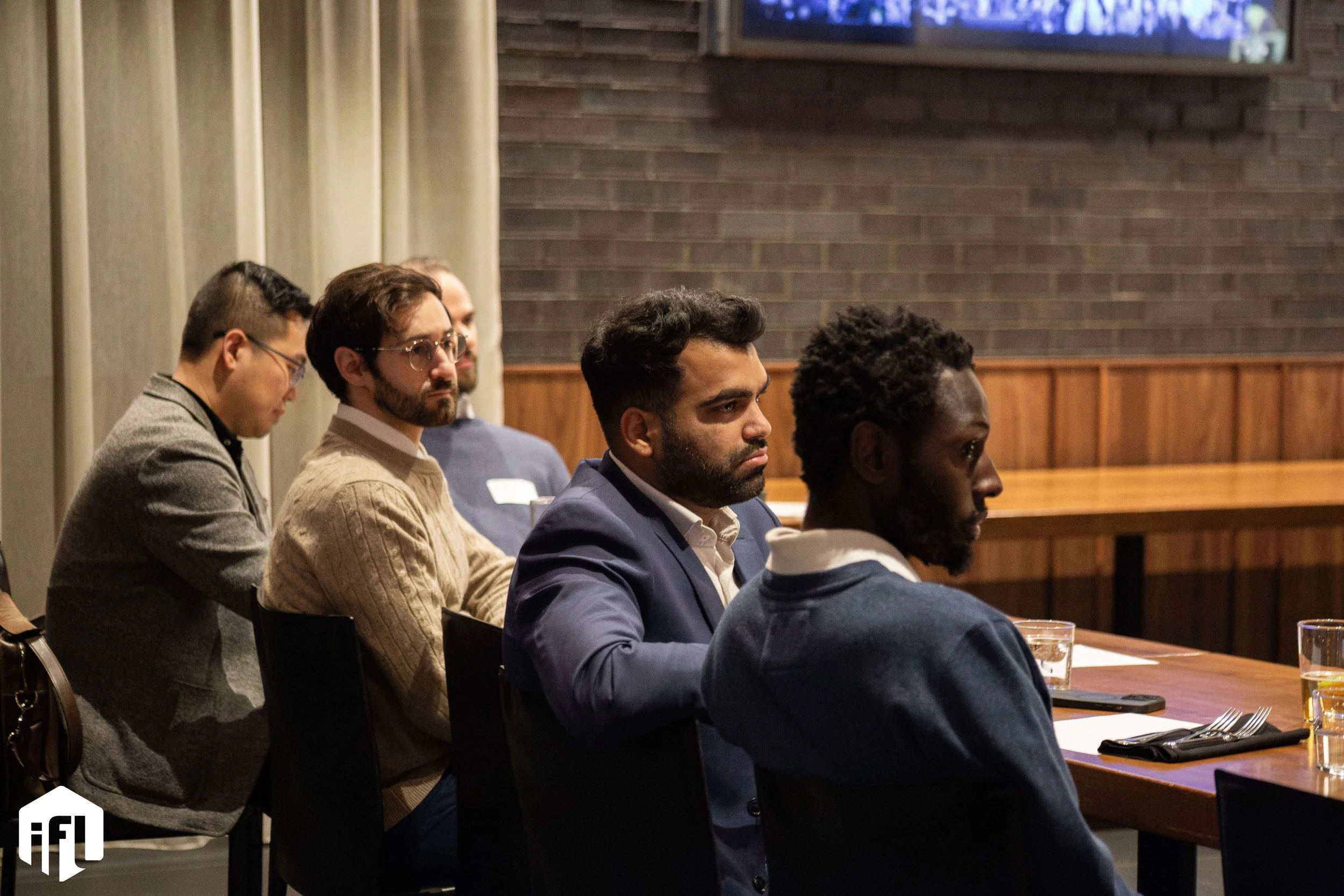 A group of people attending a meeting or conference, seated at a long table with drinks and utensils, listening intently.