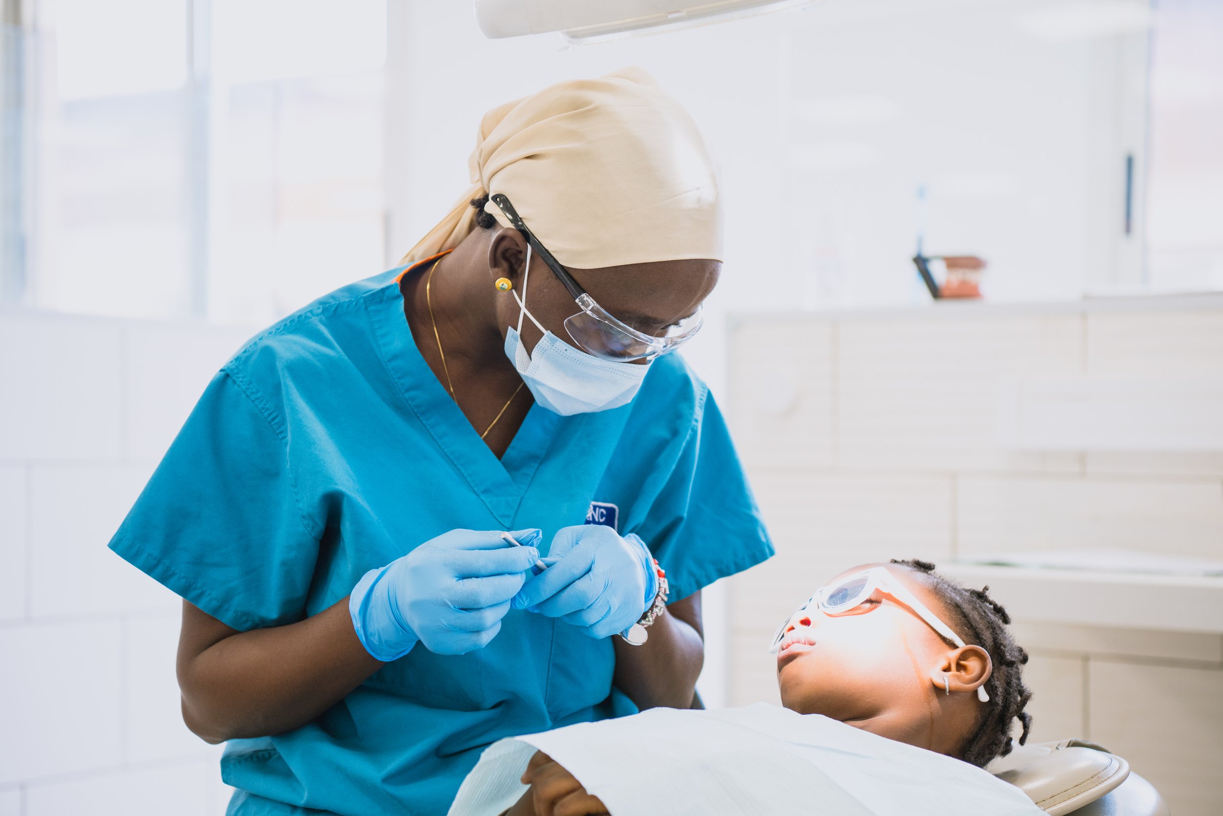 A healthcare professional, wearing blue scrubs, a beige head covering, protective eyewear, a face mask, and gloves, attending to a child patient, who is lying in a dental chair with protective eyewear, in a bright clinical setting.