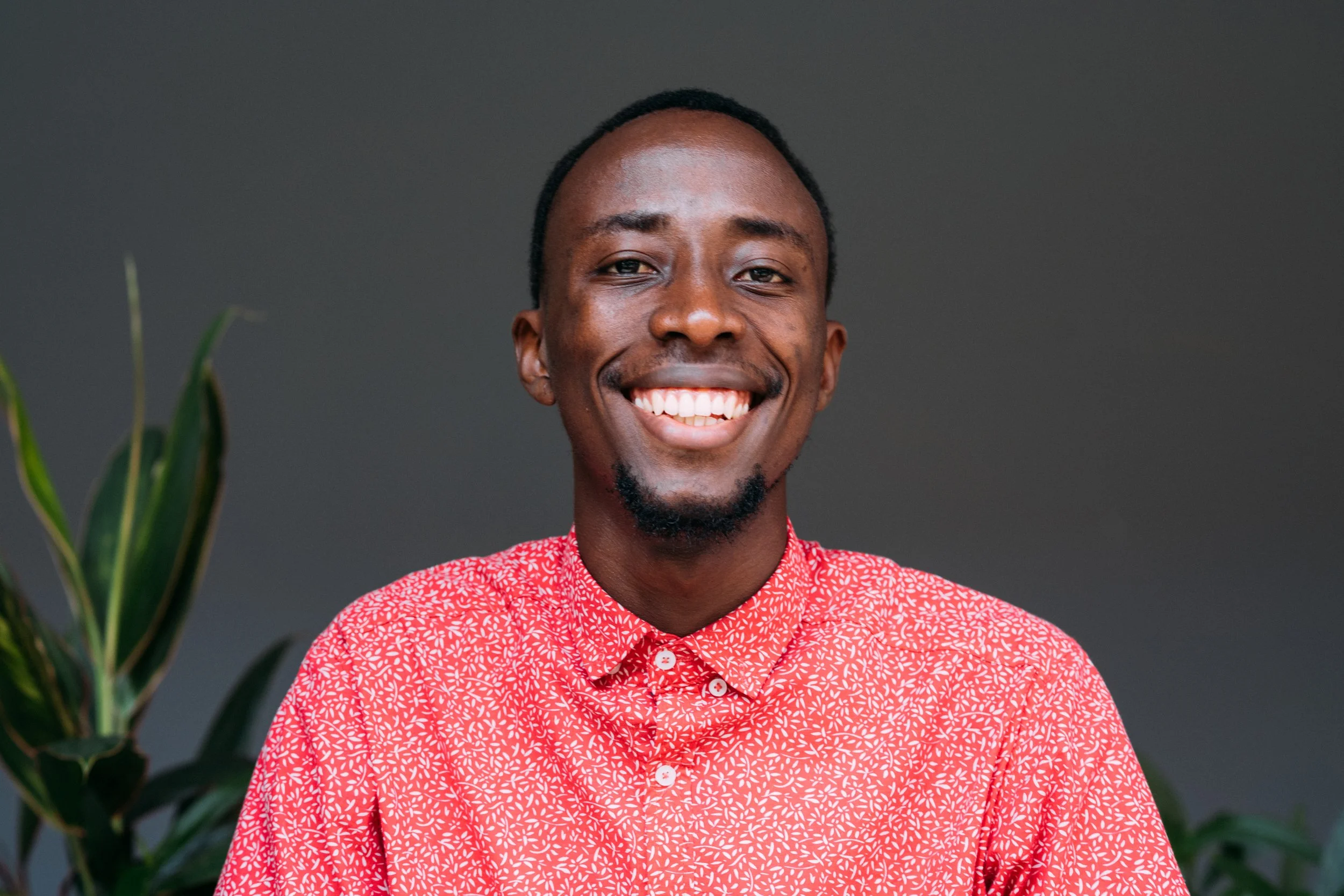 Smiling man in red patterned shirt with dark background and green plants.
