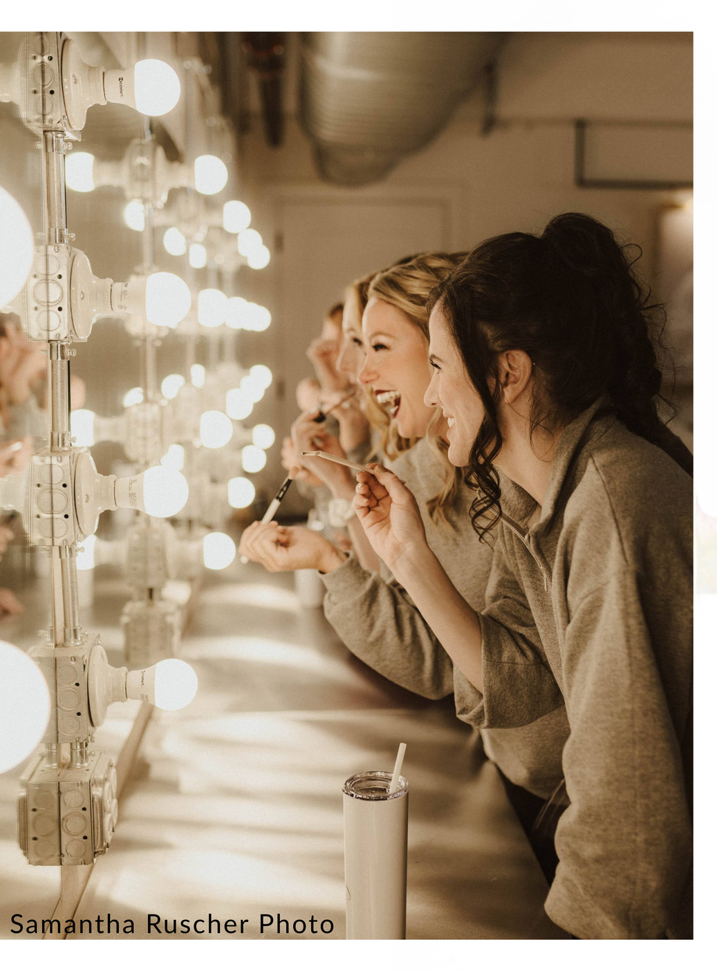 Women with makeup brushes smiling at their reflection in a brightly lit mirror at a makeup station.