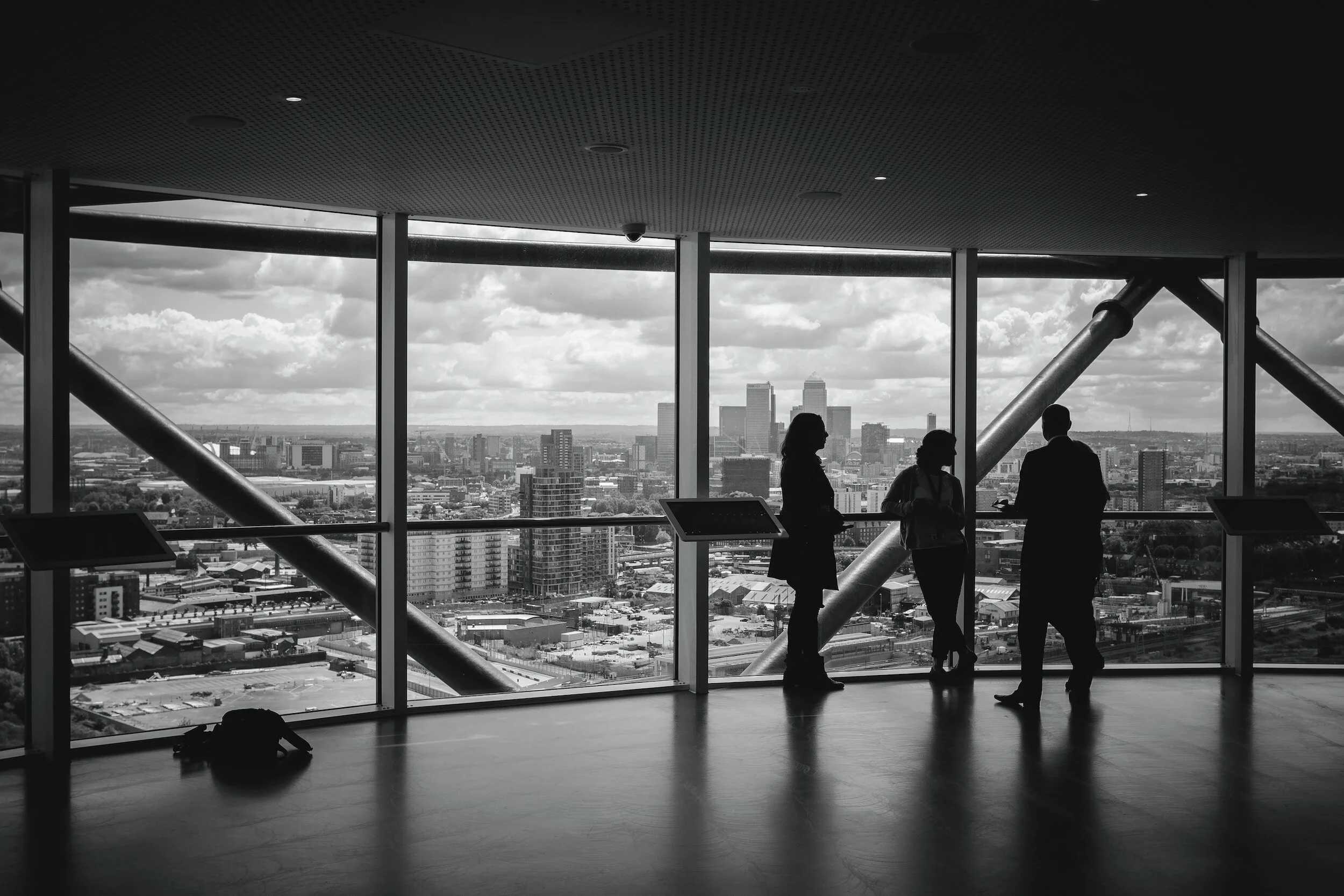 Silhouettes of three people in conversation inside a high-rise building with large windows, overlooking a cityscape with tall buildings and a cloudy sky.