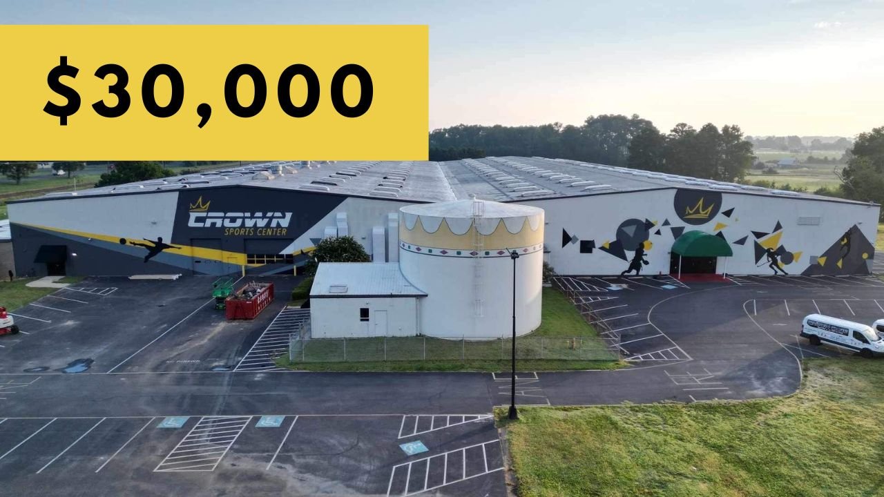 A large sports center building called 'Crown Sports Center' with a mural of athletes in action on the front, a parking lot with a few vehicles, and a yellow banner in the upper left corner displaying '$30,000'.