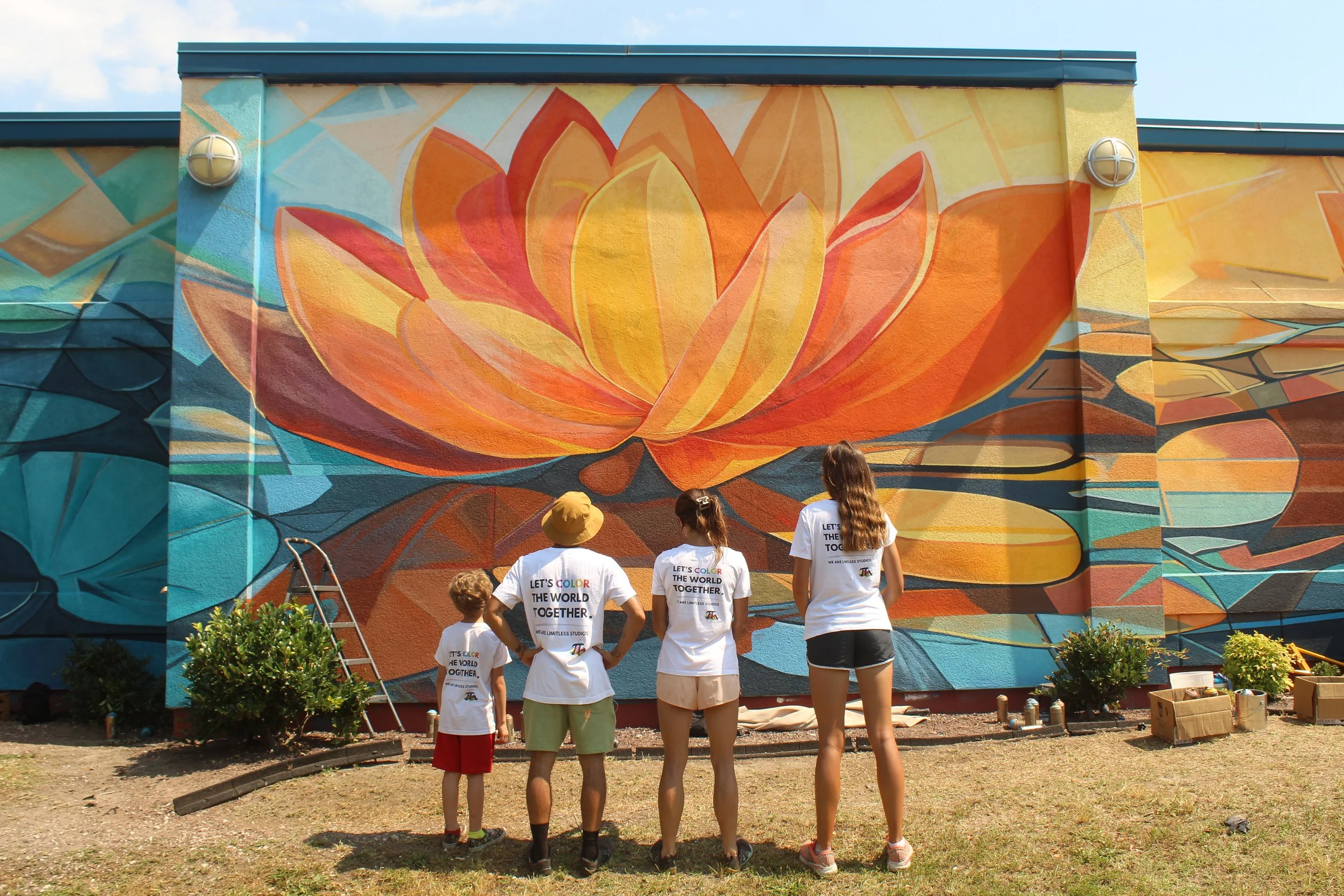 Four people stand in front of a colorful mural of a large orange and yellow lotus flower painted on a wall, with art supplies and potted plants nearby, during daytime.