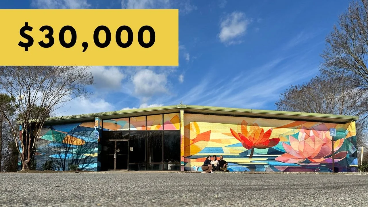 Front of a building with a colorful mural of water lilies, three people sitting in front, and a yellow sign with '$30,000' in black text.