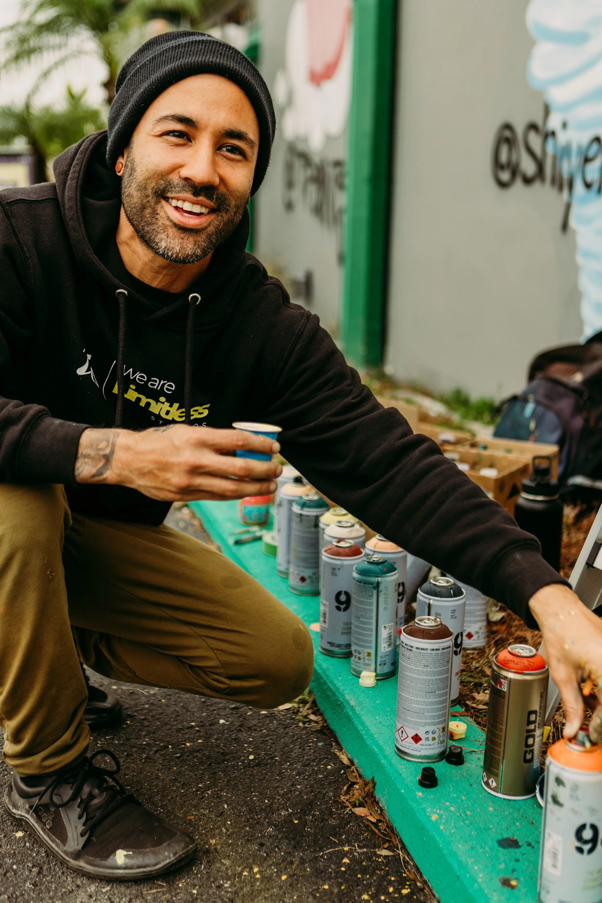 A smiling man wearing a black hoodie and beanie crouching down, holding a spray paint can, next to a row of spray paint cans on a green surface outdoors.