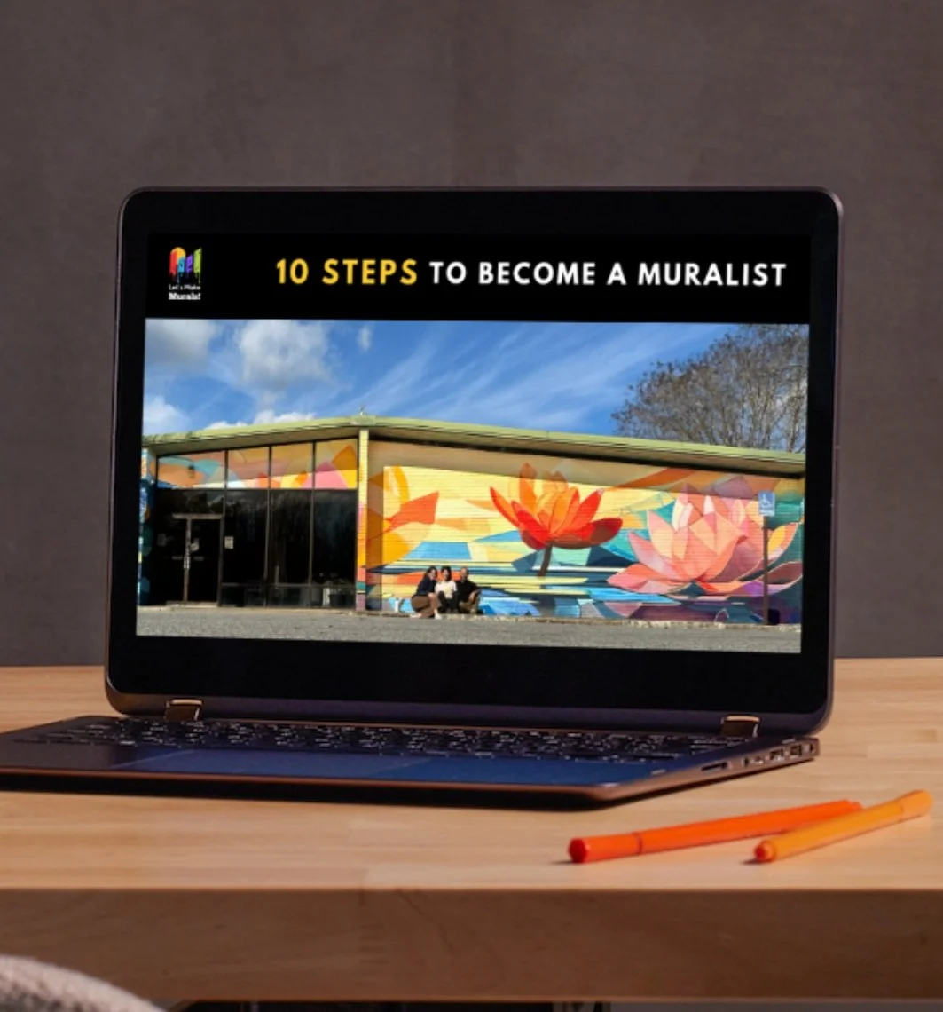 Laptop on a wooden desk displaying a mural of lotus flowers and the text '10 Steps to Become a Muralist'; orange pens on the desk in front of the laptop.