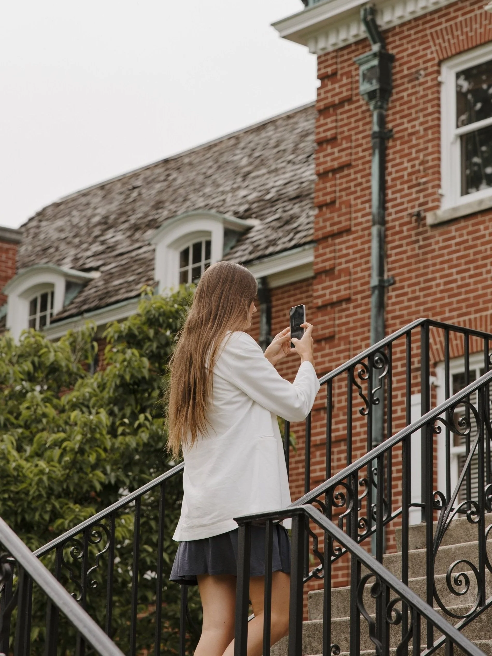 Young woman with long hair in a white jacket taking a photo with her phone on stairs outside a red brick building.