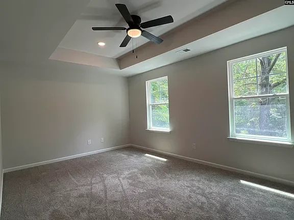 Empty room with beige carpet, white walls, two windows showing trees outside, and a ceiling fan with black blades.