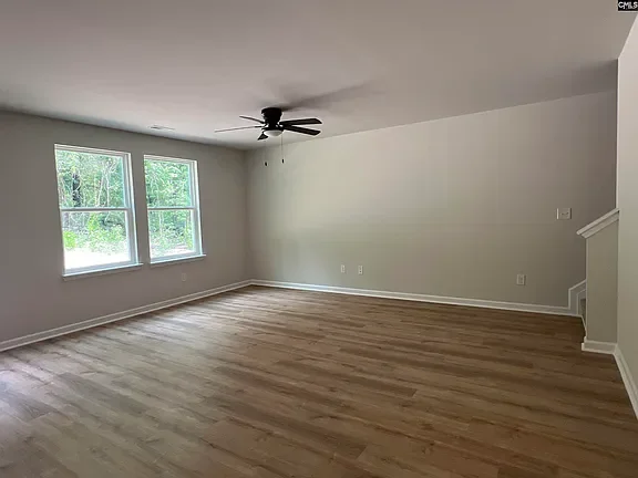 Empty living room with hardwood floors, three windows, white walls, a ceiling fan, and a staircase corner.