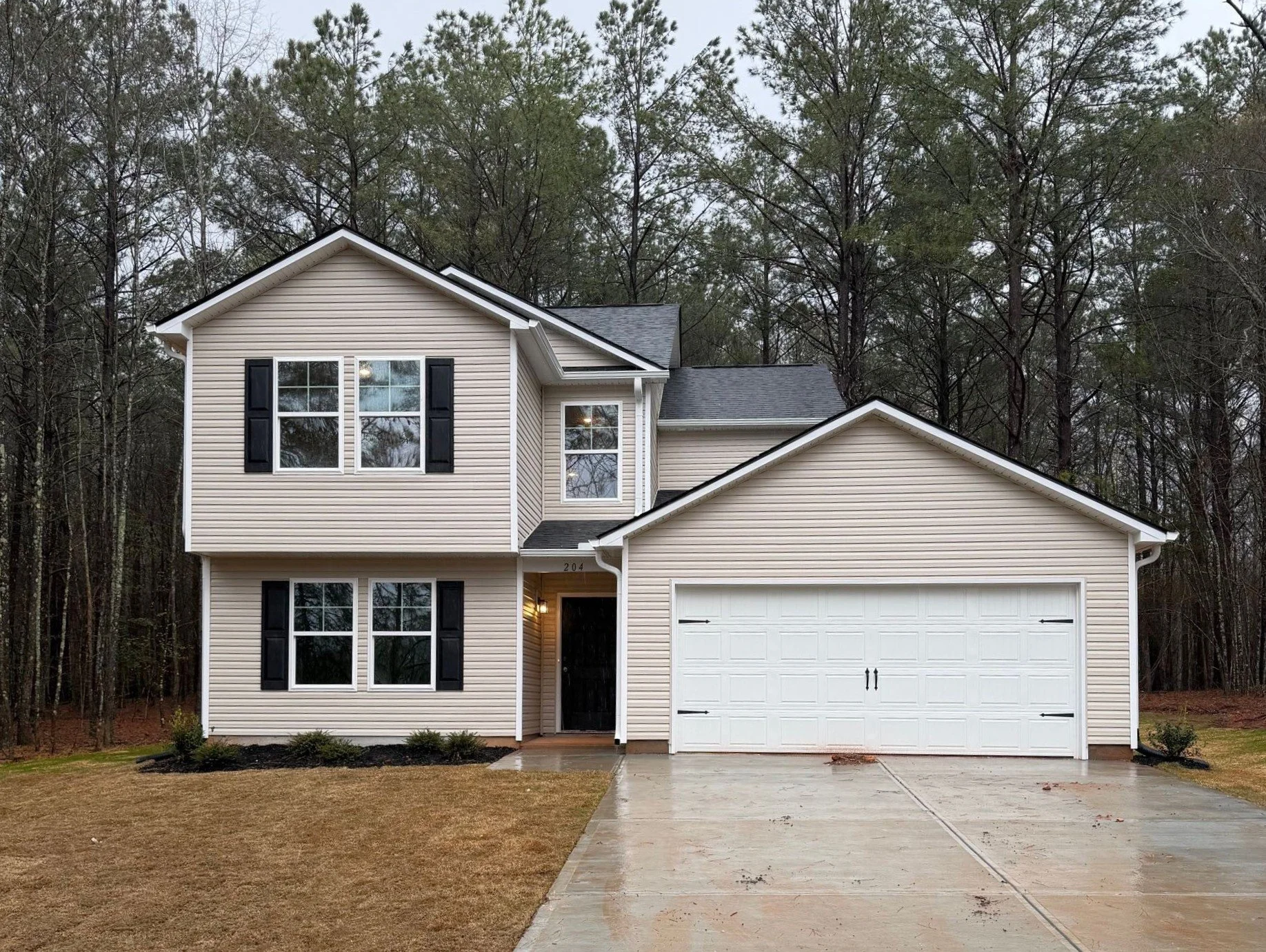 New two-story house under construction with exterior sheathing, window frames, and garage door opening visible, surrounded by a wooded area, during daytime.