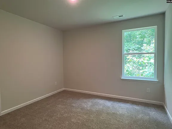 Empty room with beige carpet, white walls, a window showing green trees outside, and a ceiling vent.