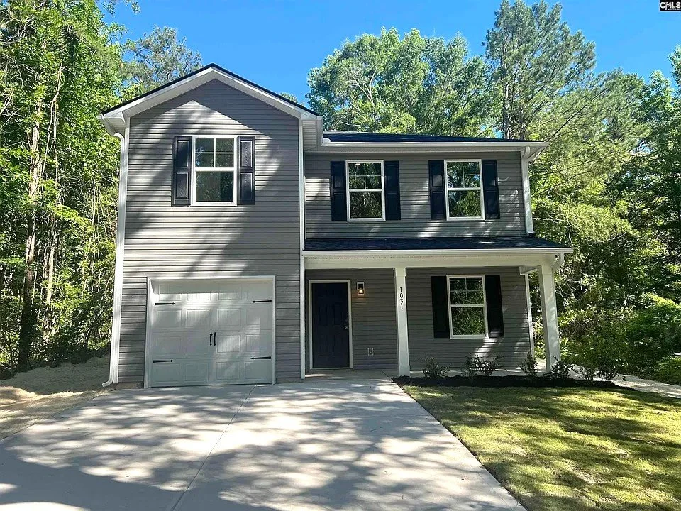 A gray two-story house with black shutters, a front porch, and a single-car garage, surrounded by trees on a sunny day.