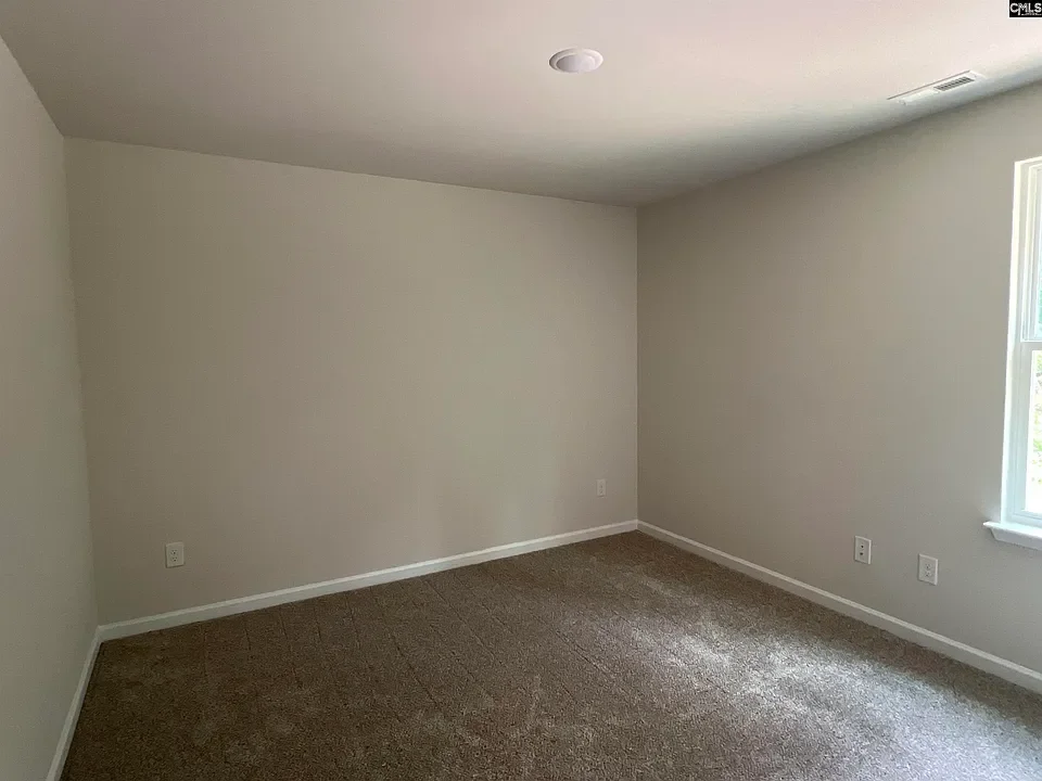 Empty room with beige walls, brown carpet, a window on the right, and no furniture.