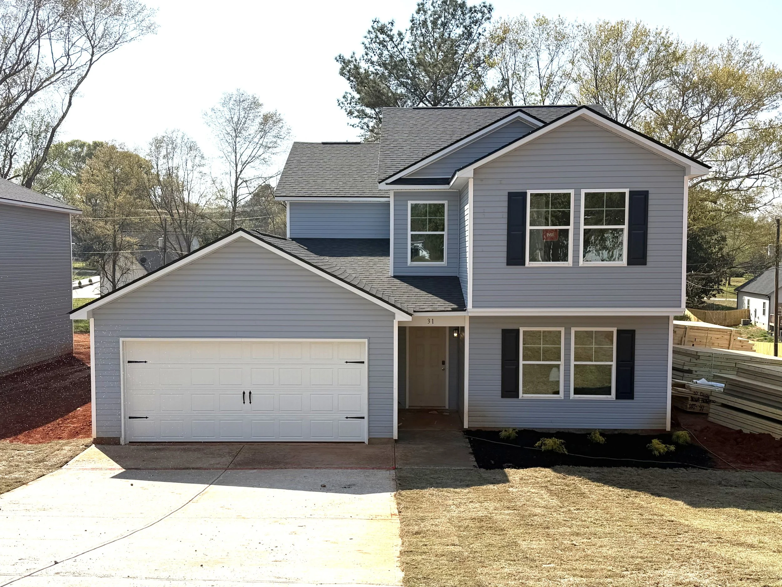 A two-story house under construction with exterior walls covered in building wrap that has blue logos, and an attached garage with an open entryway, set on a muddy yard with patches of snow.