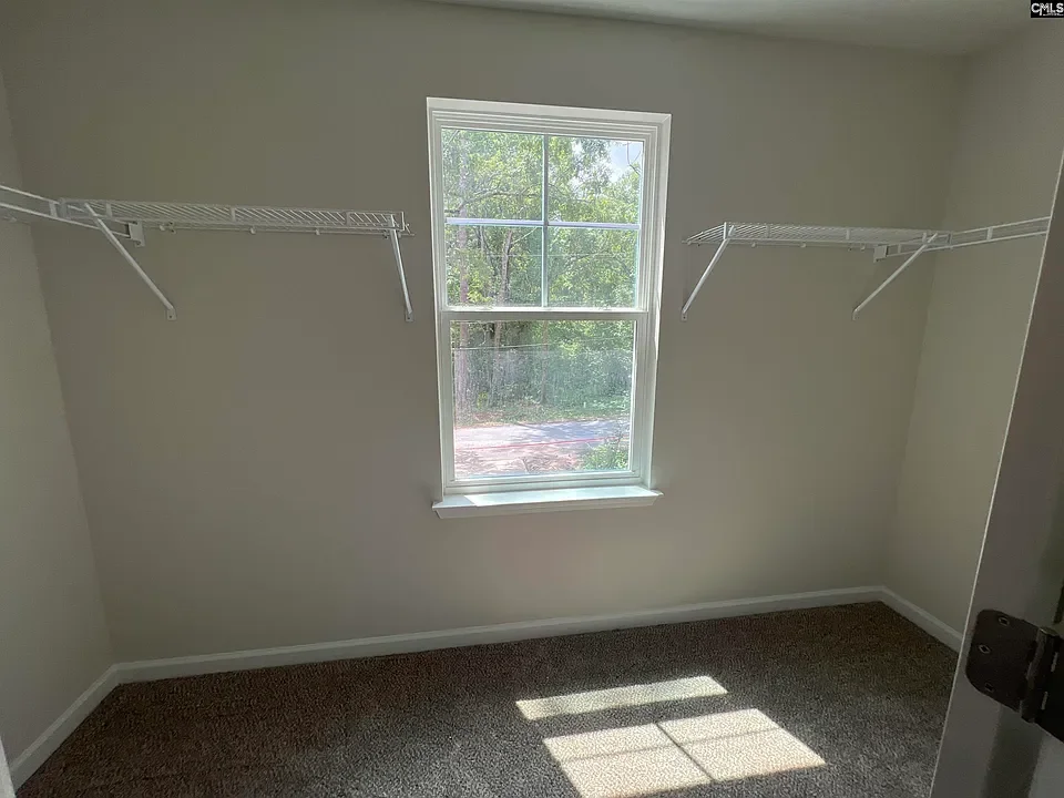 Empty room with beige walls, a single window, and white wire shelves on two sides, with brown carpet flooring.