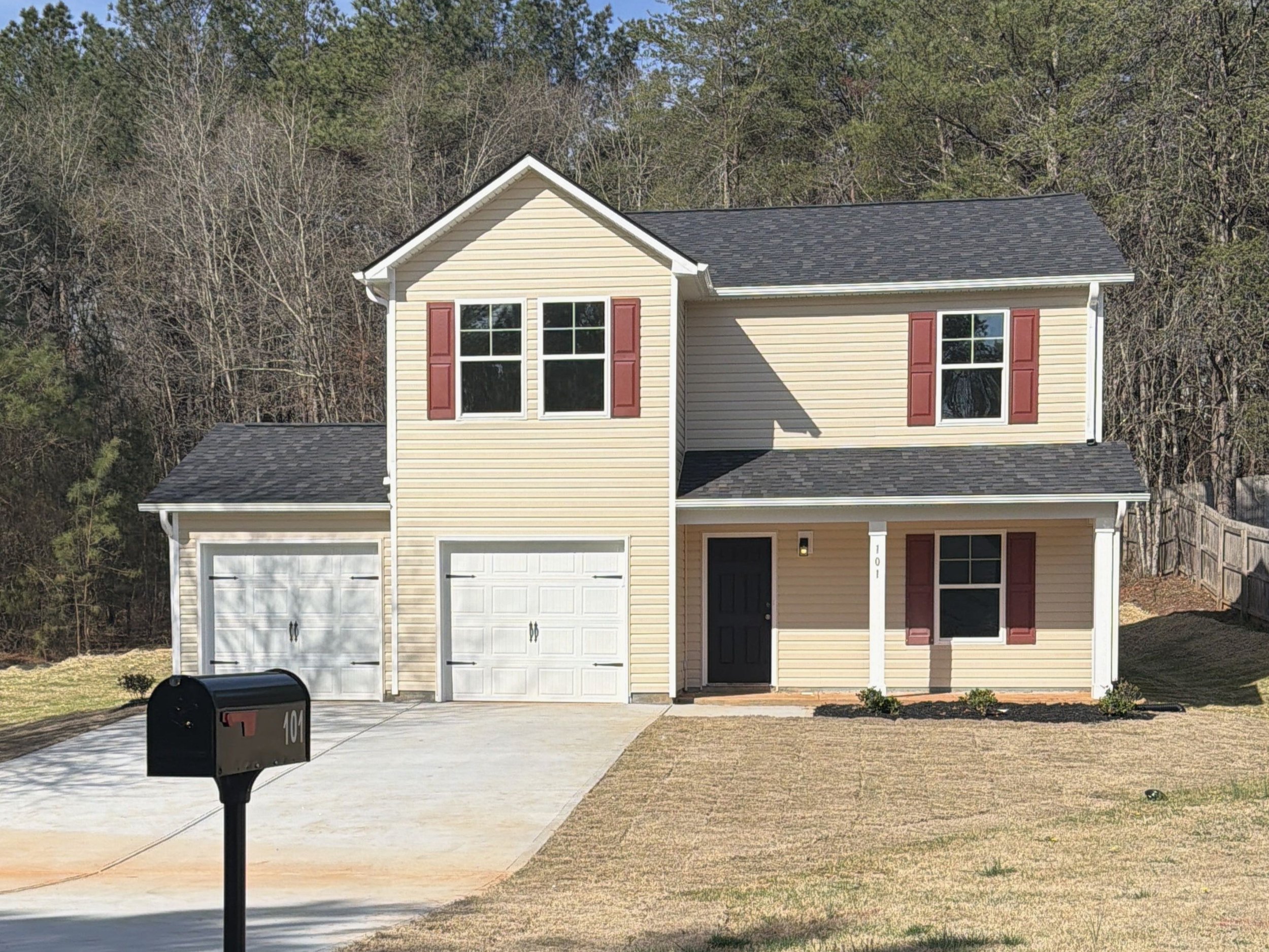 Under construction two-story house with exposed wood framing, windows installed, and a sloped roof. Construction materials and tools are on the ground in front of the house. The yard is undeveloped with dirt and some grass, surrounded by trees and a wooden fence on the right.