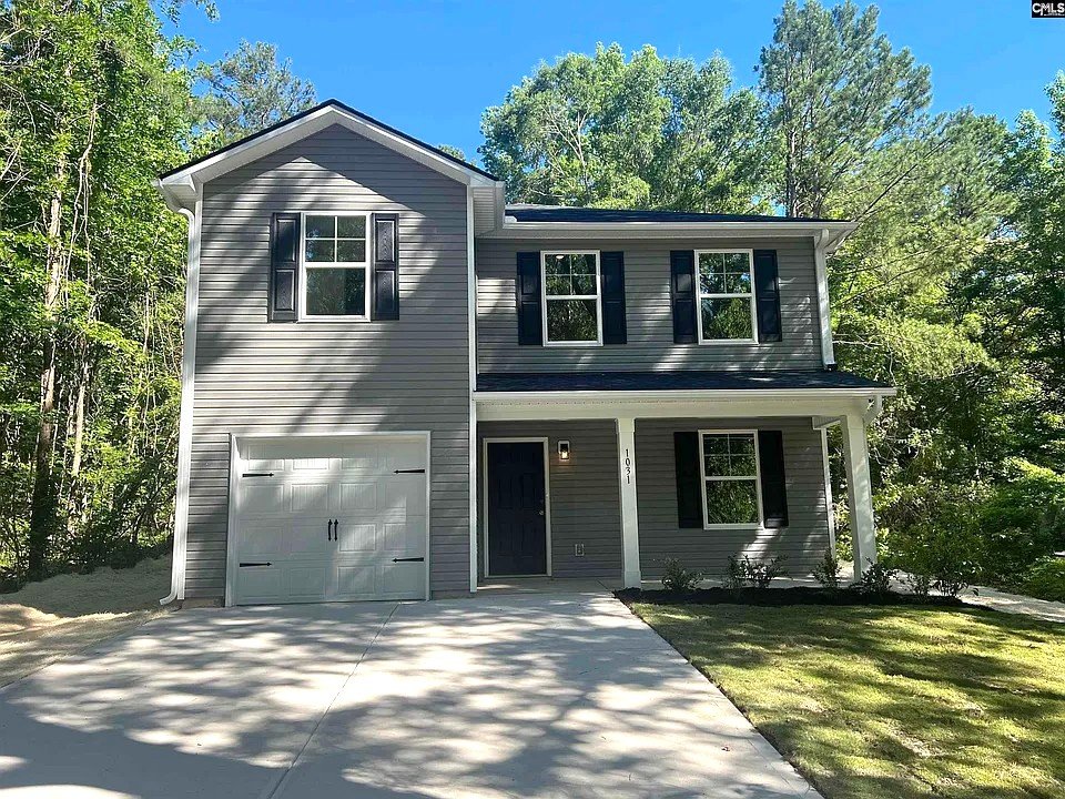 A two-story gray house with black shutters, a garage, and a front porch, surrounded by trees, under a clear blue sky.
