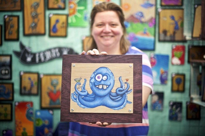 Katie Call from Artsplosion holding wooden frame with 2D graphic art of a blue octopus drinking a martini.
