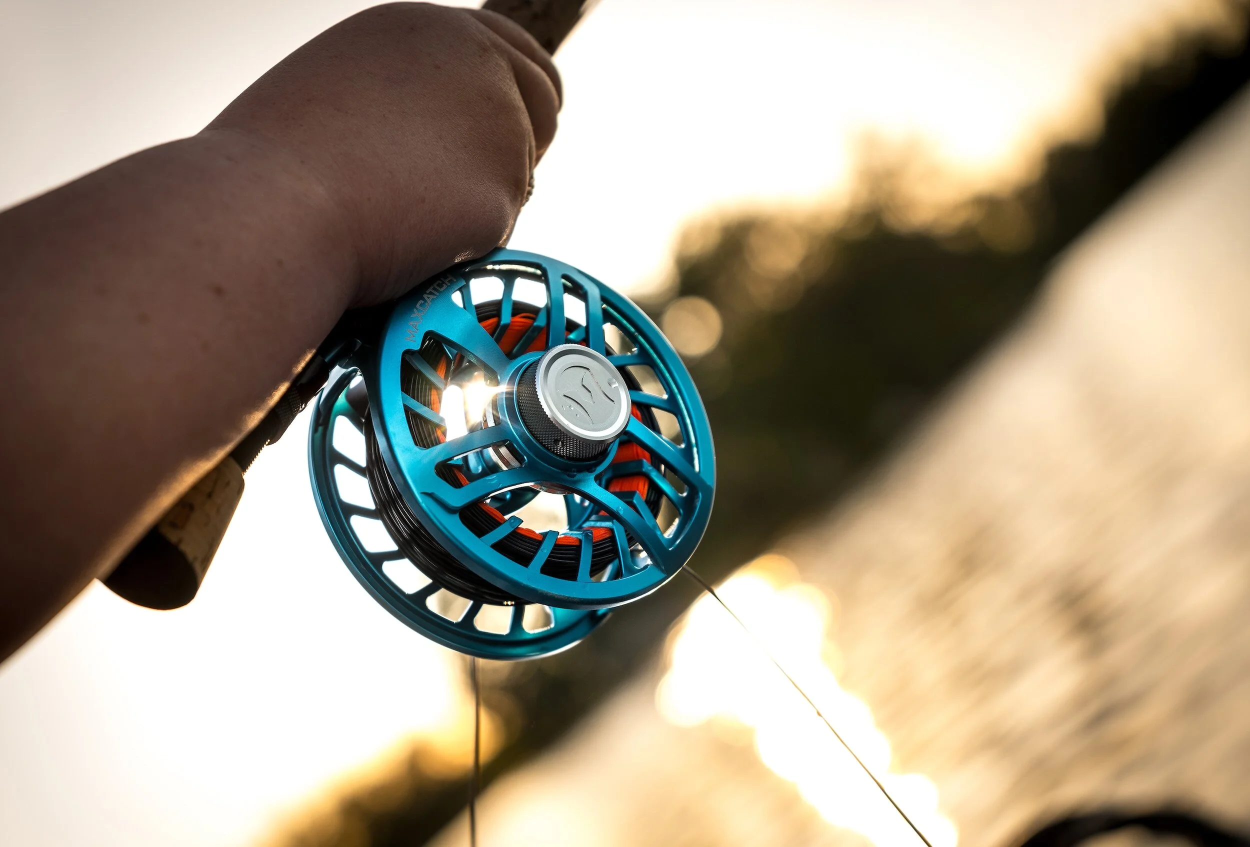 A person holding a blue fly fishing reel with orange internal components, shown against a sunset background by a body of water.