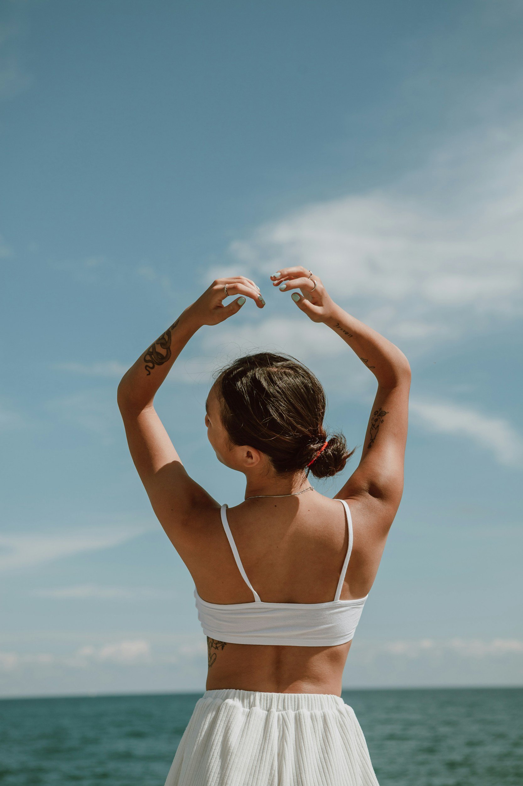 Jeune femme avec des tatouages, portant un haut blanc et une jupe blanche, se tenant à la plage avec la mer en arrière-plan, levant les bras au-dessus de la tête contre un ciel bleu.