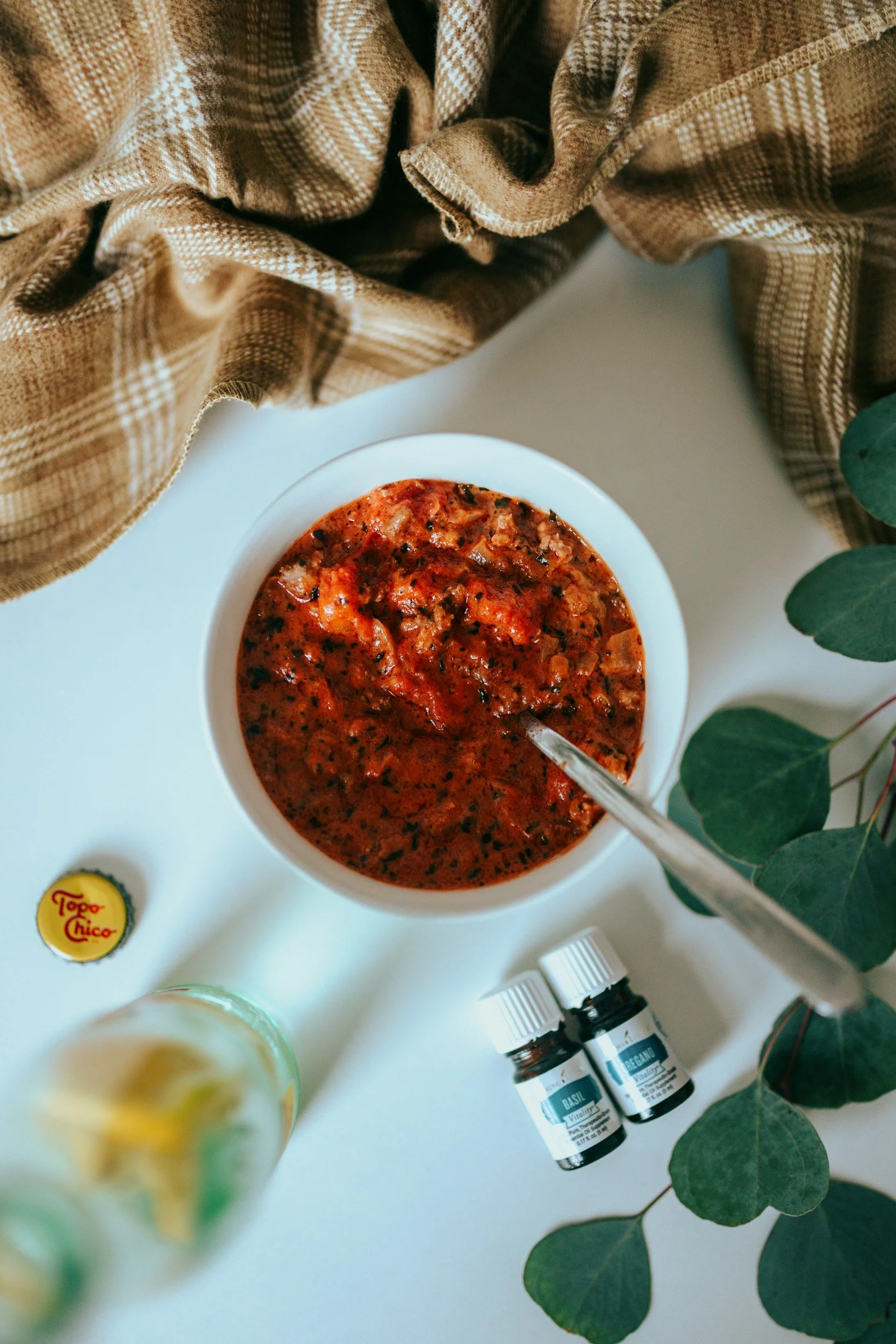 Soupe tomate dans un bol blanc avec des bouteilles d'huiles essentielles à proximité, une étiquette de conserve Topo Chico, et des feuilles vertes de eucalyptus sur une surface blanche, avec un tissu en laine beige à l'arrière-plan.